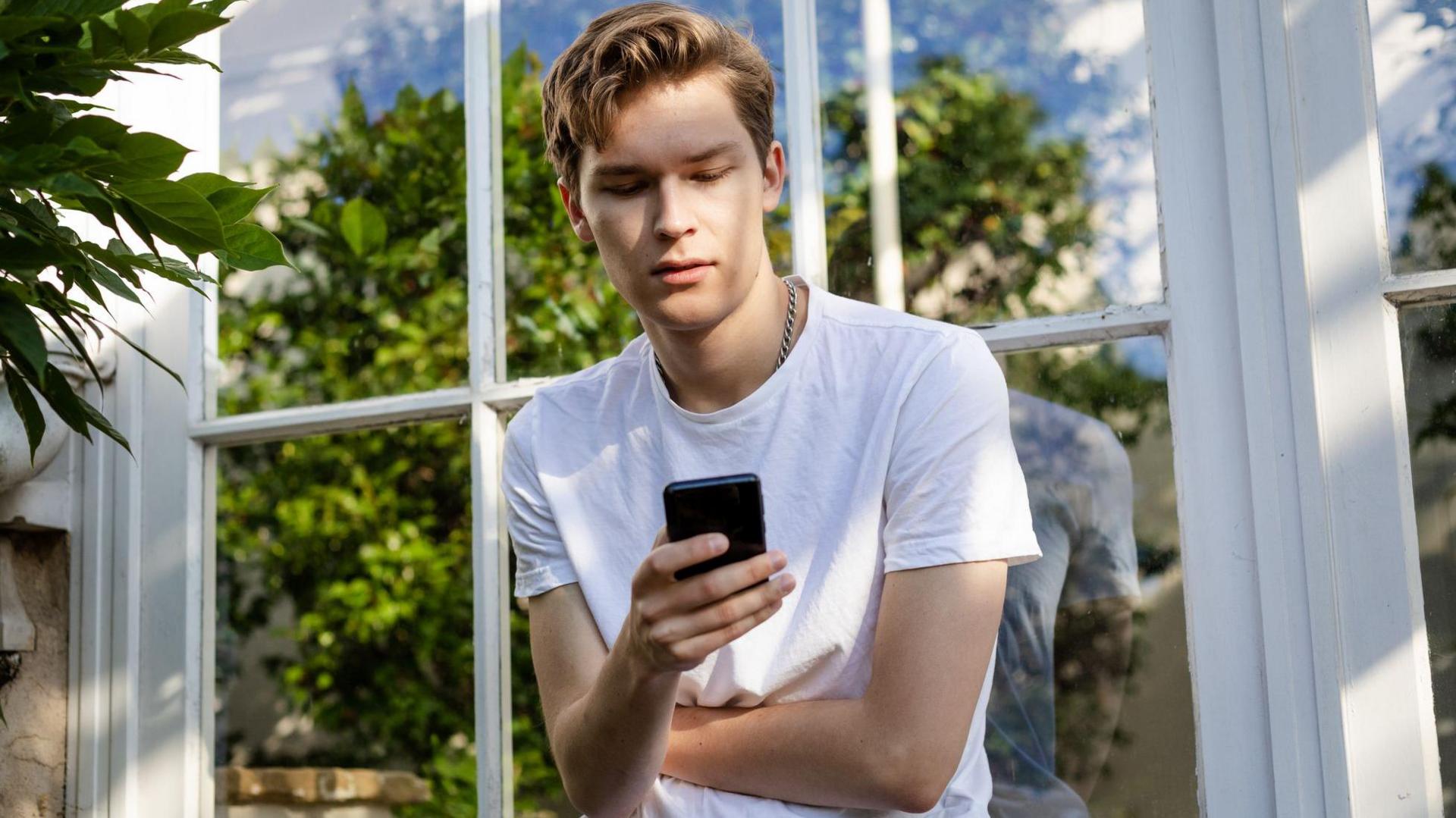 A young man stands outside a house looking at his phone with a reflection of a tree in the window behind him