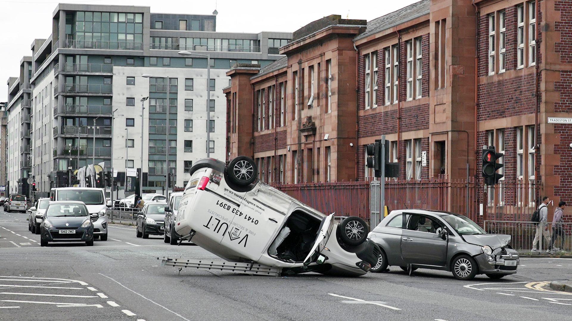 A white van with a ladder on top flipped on it roof with a small grey car with a smashed bonnet beside it.
