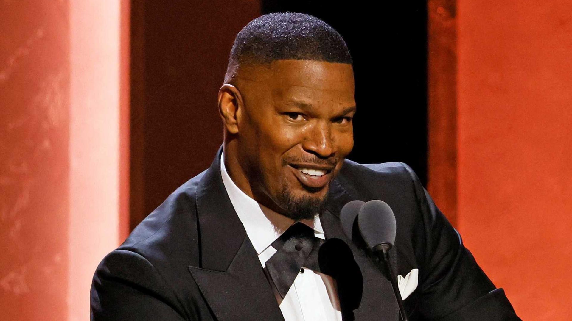 Jamie Foxx is wearing a black tuxedo with a white shirt and is smiling at the camera