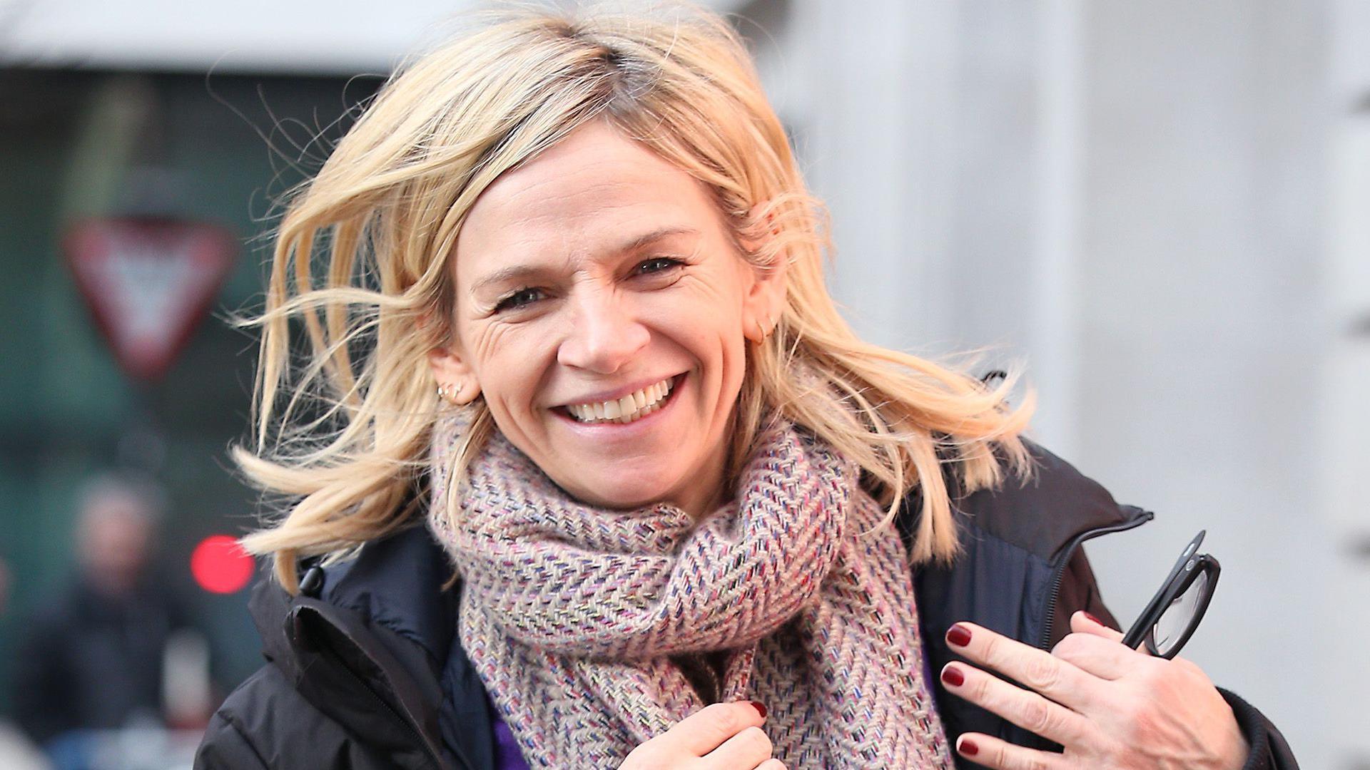 Zoe Ball smiling in a black jacket and brown scarf.