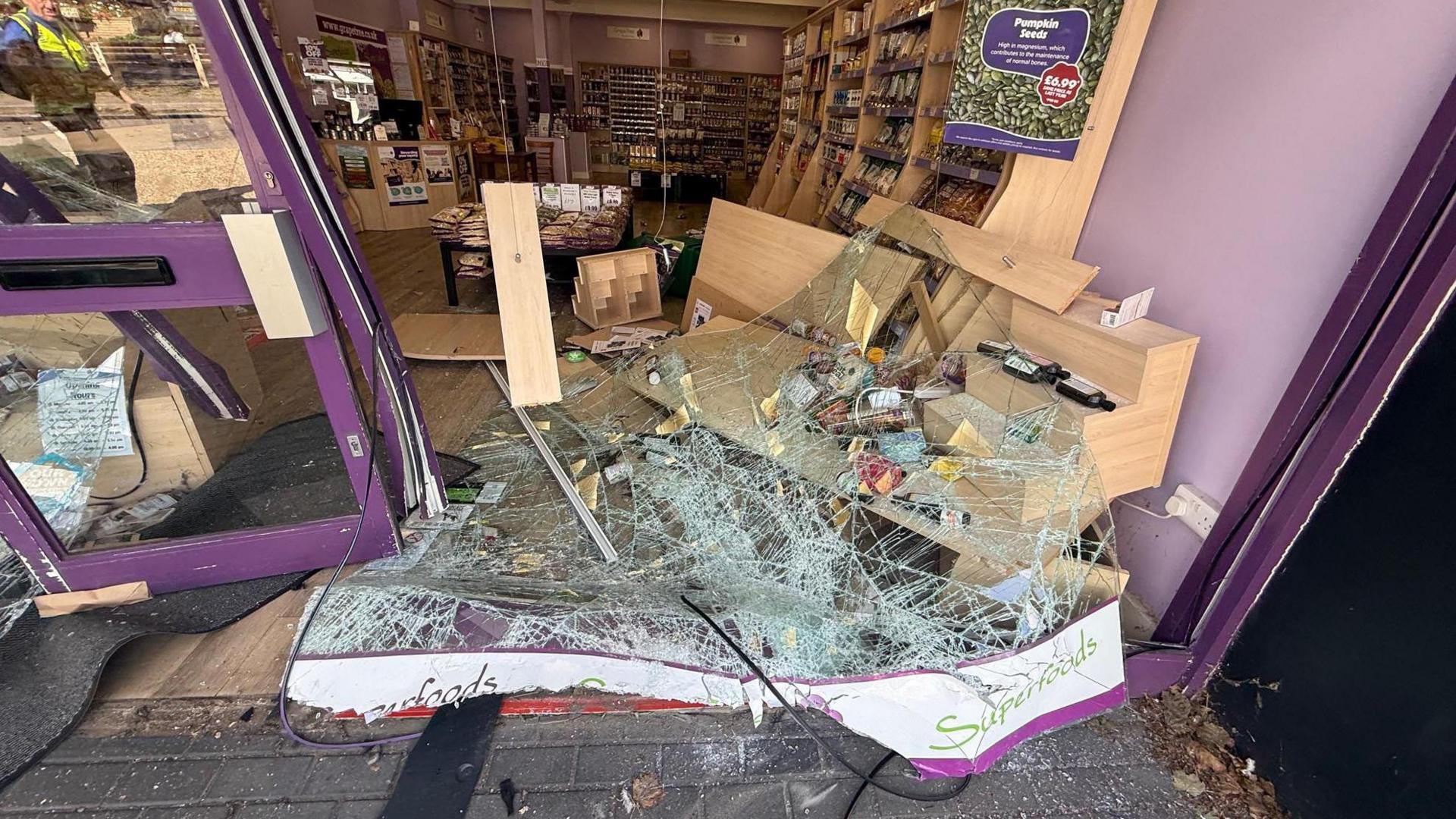 The picture shows the front of a store that has been destroyed. There is broken glass on the floor, the doors are bent inwards, shelves have fallen along with the goods in them. The back of the store is completely untouched, as products can be seen lined up neatly on shelves.