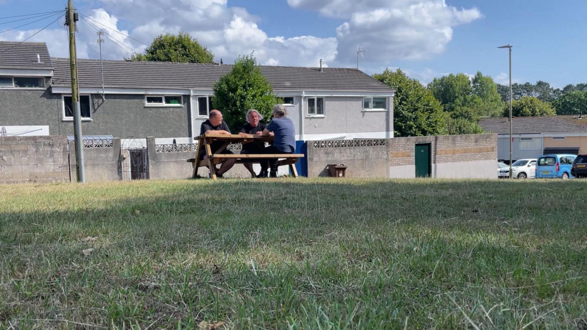 Three men sit on a park bench on Wilmot Gardens in Plymouth. Several terrace houses with grey walls are behind them along with a few trees.