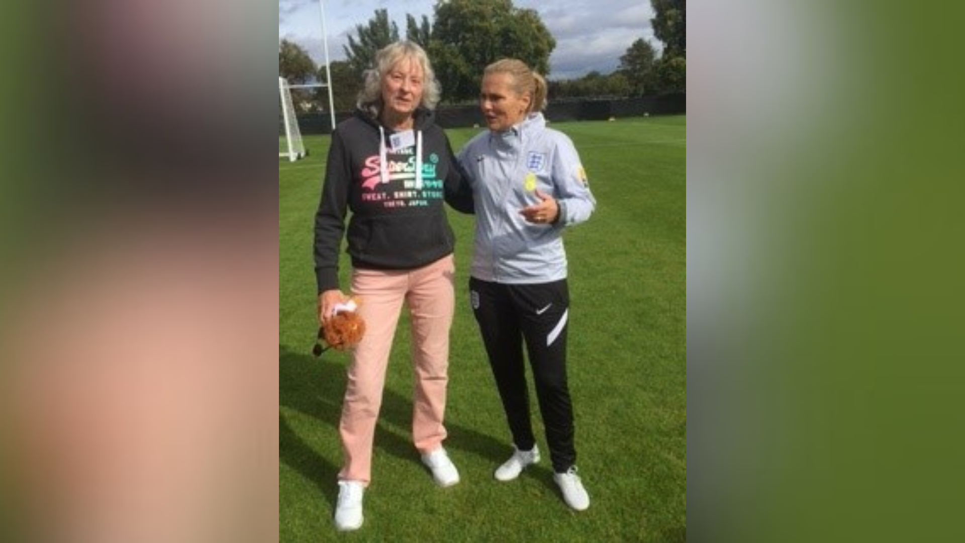 Two women stood next to each other on a football pitch. One is wearing a black hoodie and salmon pink trousers. The other is wearing a grey tracksuit top and tracksuit trousers.
