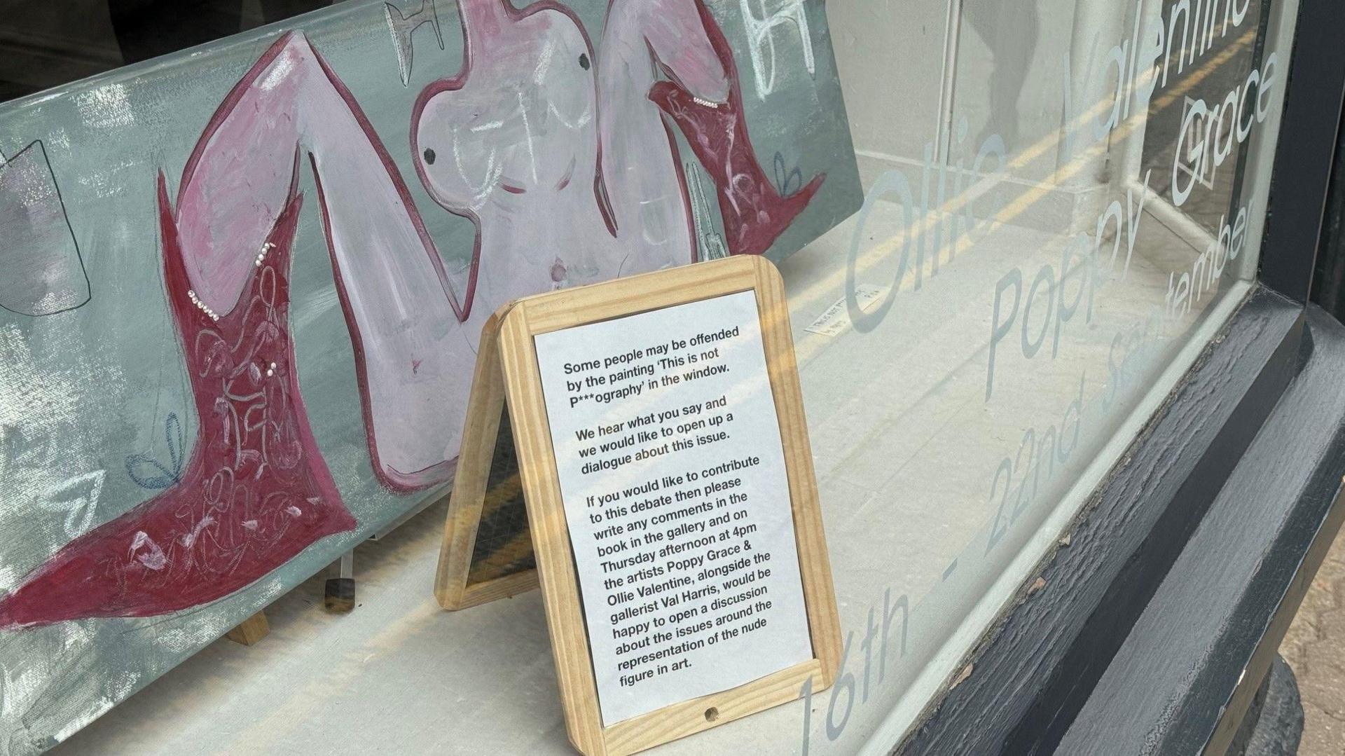 A painting of a naked woman with her legs spread in a gallery window, with a small notice in front of the painting telling visitors they could leave comments in a book. 