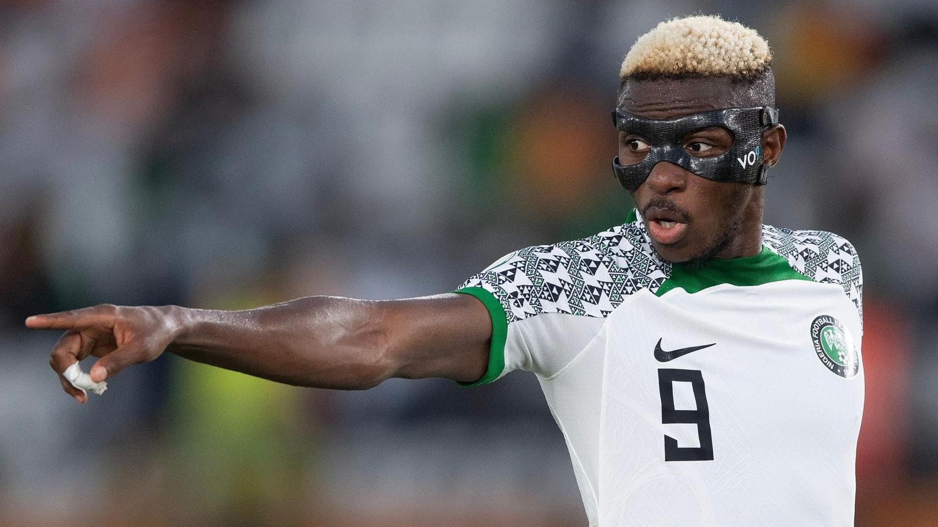 Victor Osimhen, wearing a white Nigeria shirt and a black protective face mask, points with his right arm during a football match