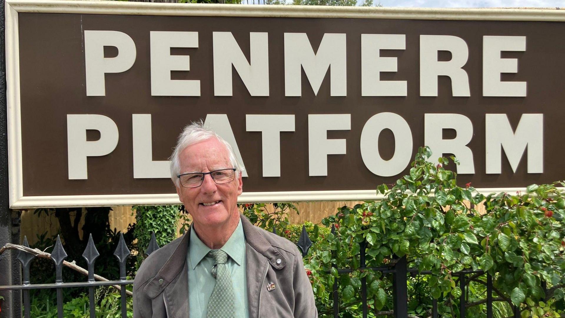 A man standing in front of a vintage style railway station sign that reads Penmere Platform. He has white hair and wears a green tie 