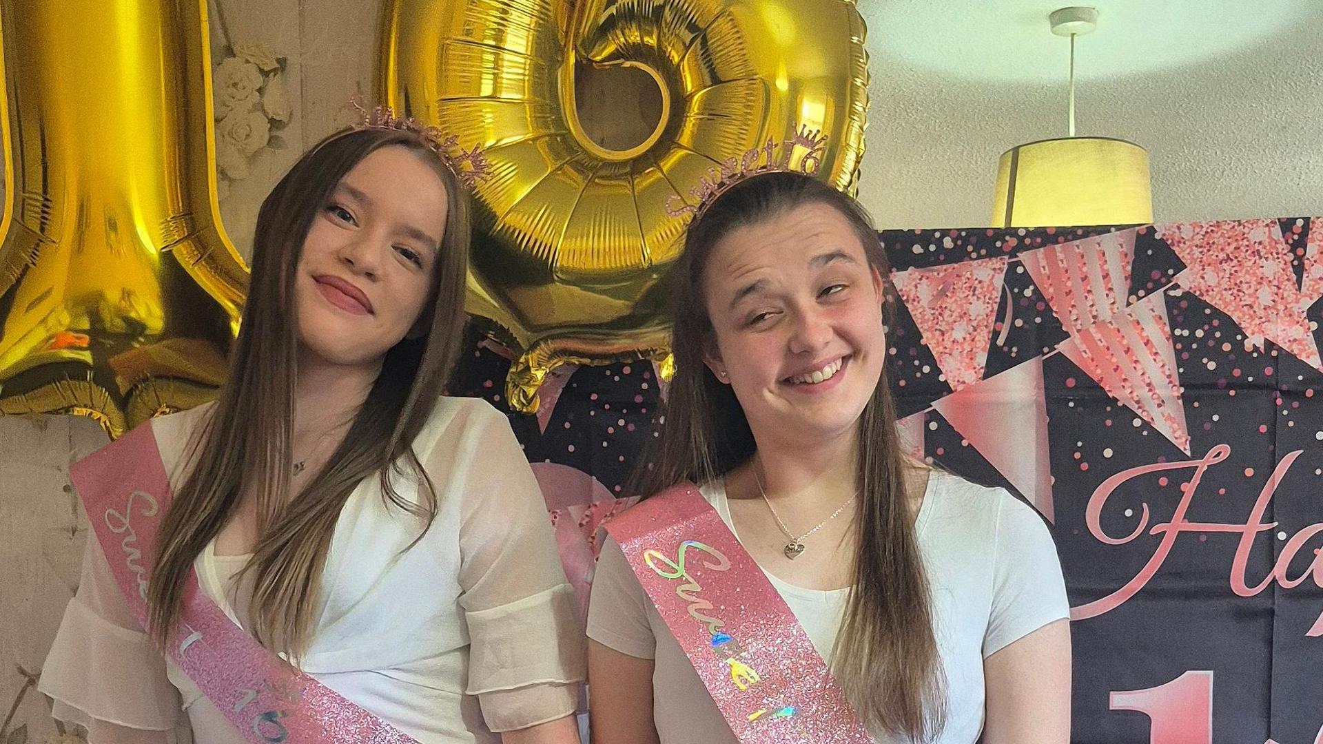 Twin sisters Jasmine (left) with long brown hair wearing a white chiffon top and Emily wearing a white t-shirt at their 16th birthday party. They both have pink sashes on to celebrate and tiaras on their head. 