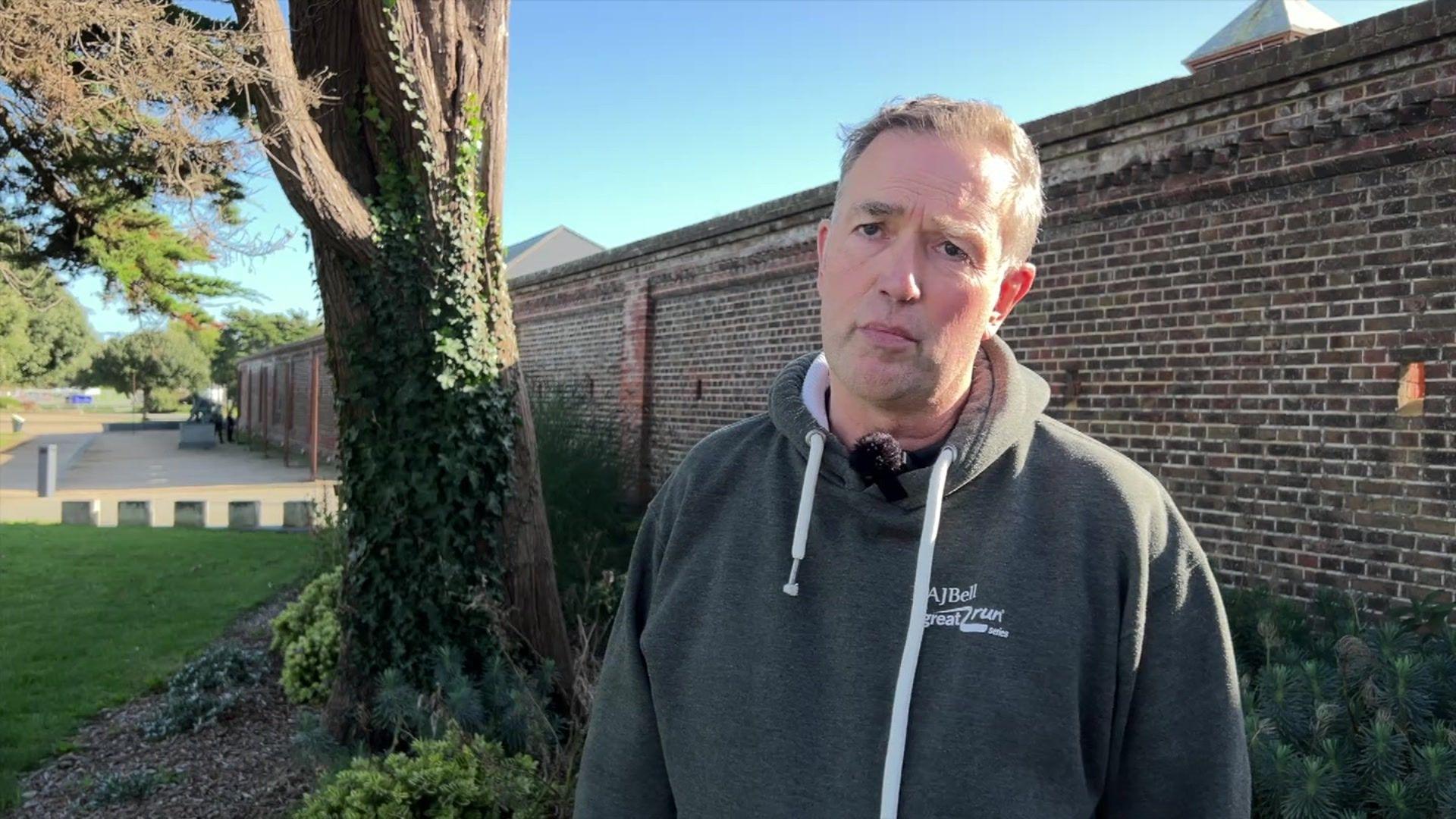 Operations director Neil Gough being interviewed after news of the event's cancellation. He is wearing a green switcher with the event's branding. He is outside, by a wall. There is a tree behind him.