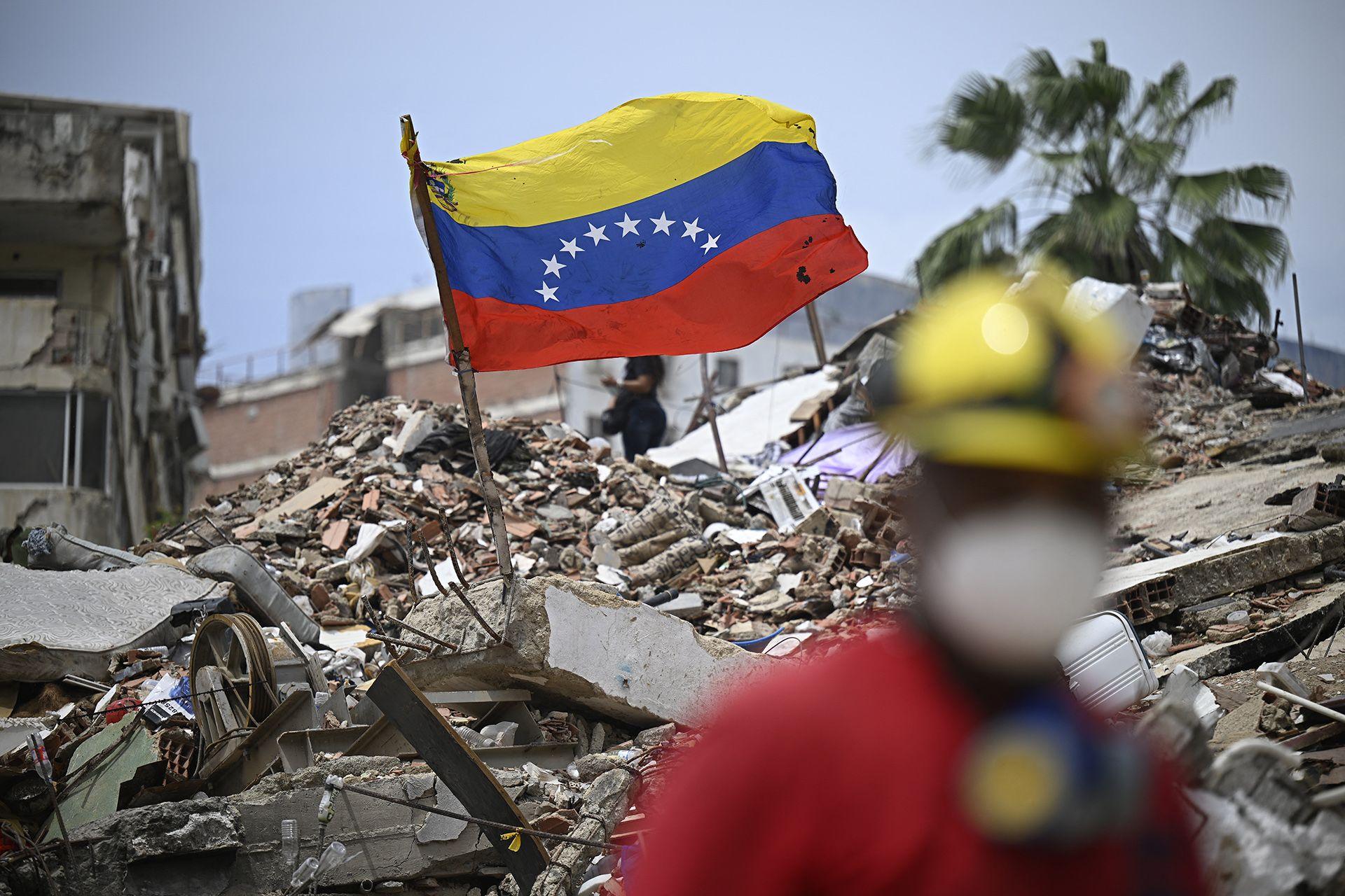 A Venezuelan flag is pictured on top of a pile of rubble with a palm tree in the background against a blue sky, while a rescue worker walks in the foreground with bokeh, in Caraballeda, Venezuela on 20 July.