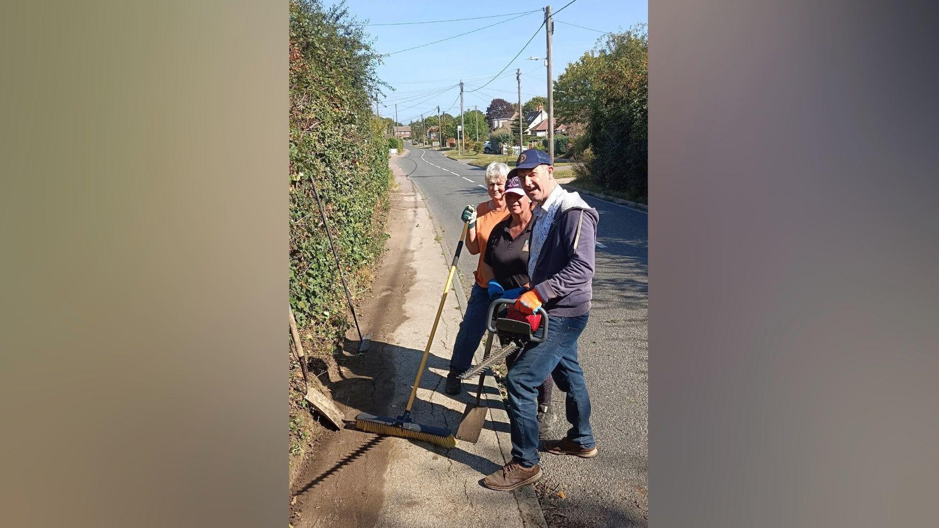 Two women and a man with gloves, hats and tools in hand, standing next to a path that has recently been cleared of weeds