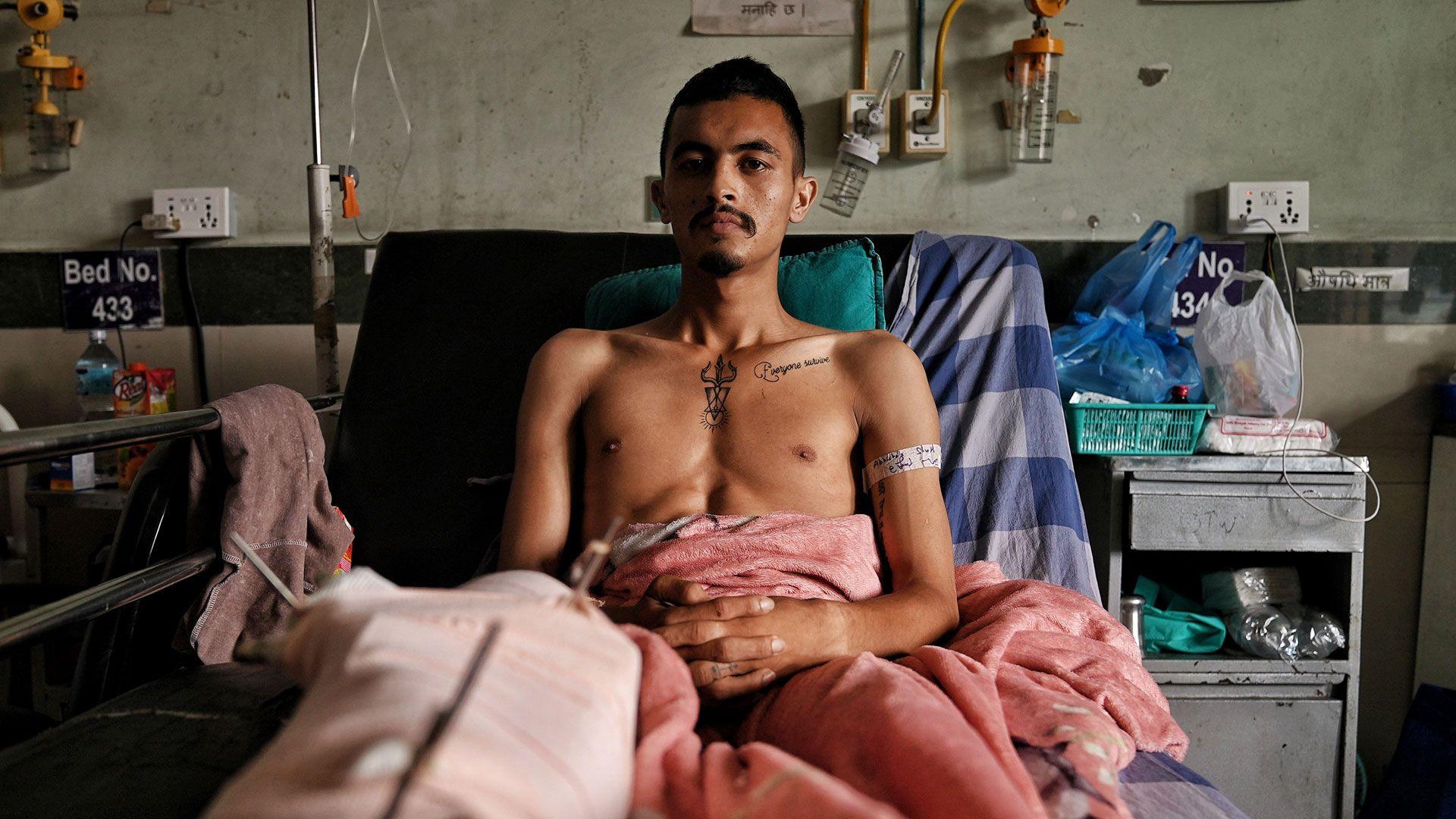 A man sits in a hospital bed, his leg in plaster. He is under a pink blanket. He has tattoos and a beard. An iron chest sits beside him with what appears to be medical supplies