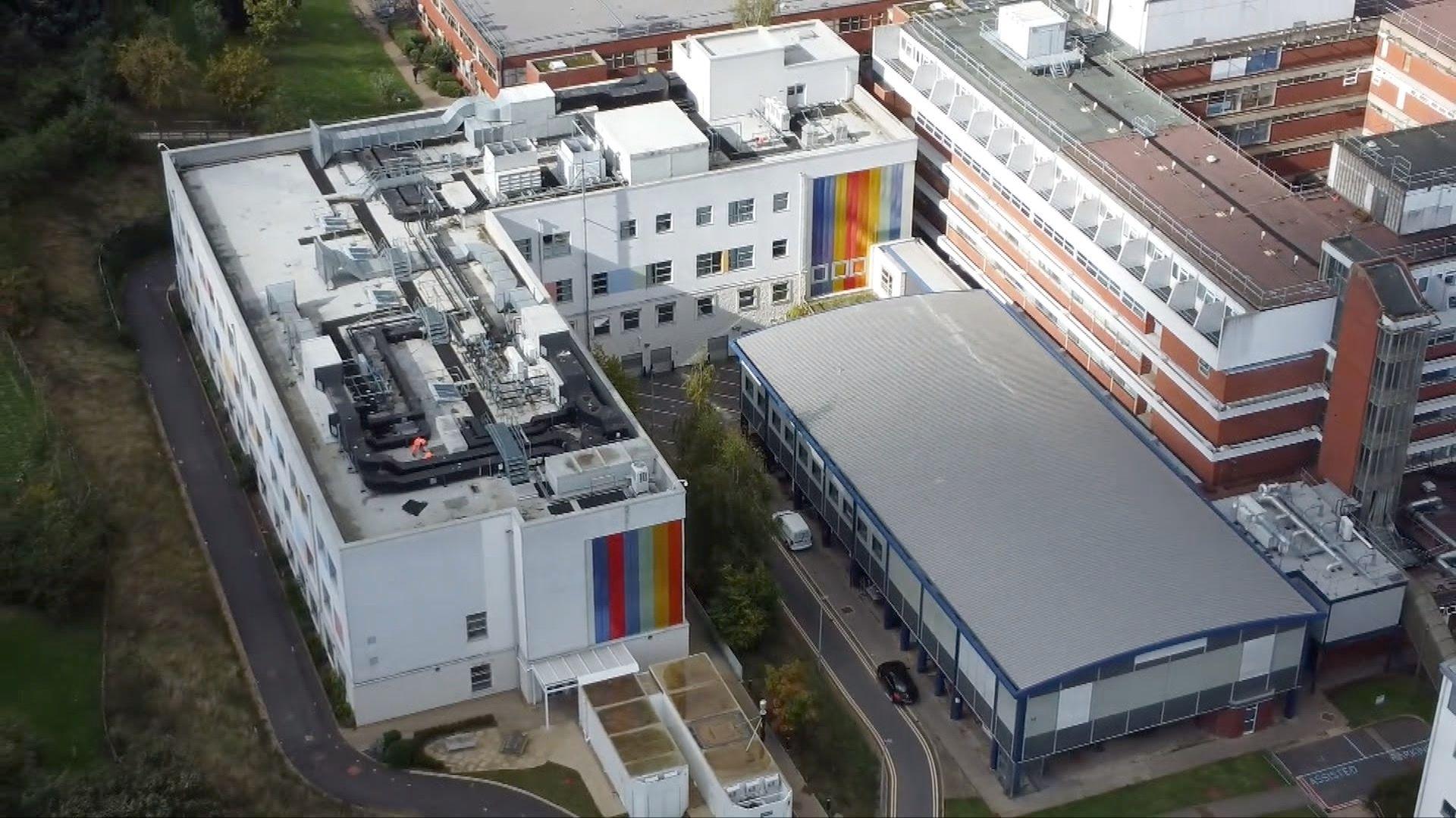 The children's ward at Kettering General Hospital from the air. The picture shows a white building on the side of the main hospital with rainbow strips on the cladding. The hospital has a flat roof.