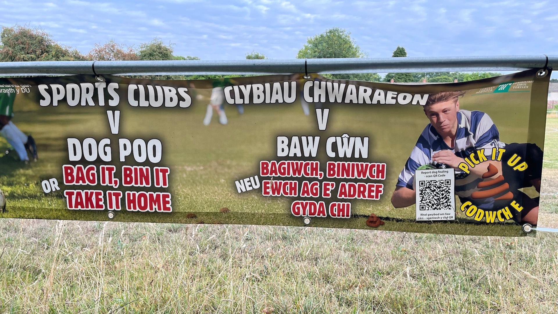 Banner in Croesyceiliog warning people against leaving dog poo. It has an image of a young boy in a blue striped shirt running with a rugby ball on it and says in England and Welsh: Sports clubs v dog poo, bag it, bin it, or take it home. 