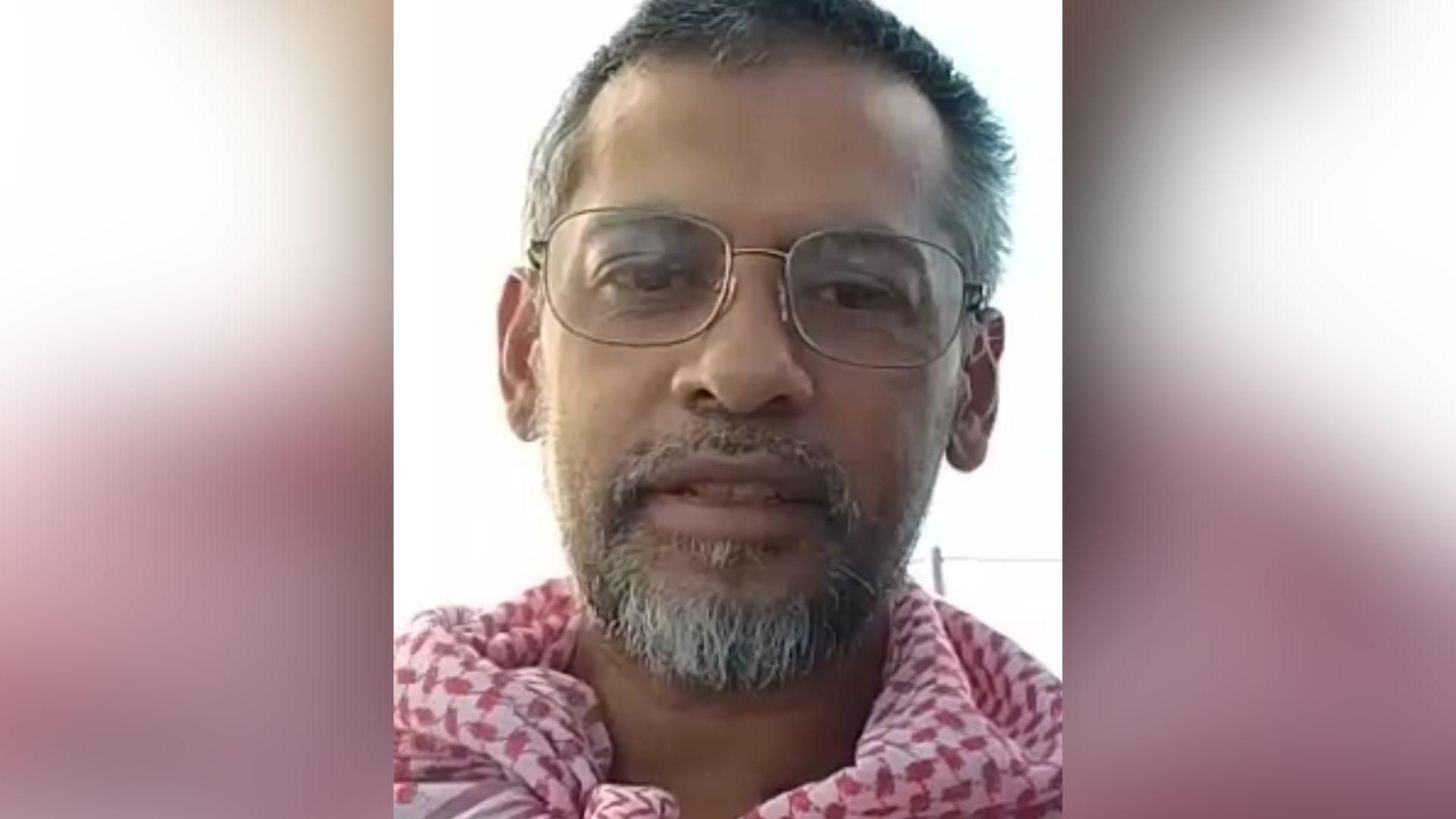 Mohammed Mokuzzal Ali. He has short dark and grey hair and facial hair. He is wearing a pair of reading glasses and a scarf with a red and white pattern. His mouth is slightly open. He is looking at the camera.