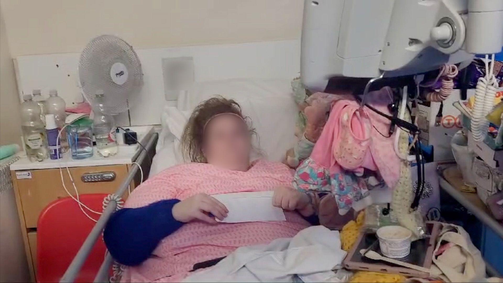 Jessie wearing a pink top and holding a letter lies in her hospital bed, surrounded by all her possessions, some of which are hanging from the monitor. 