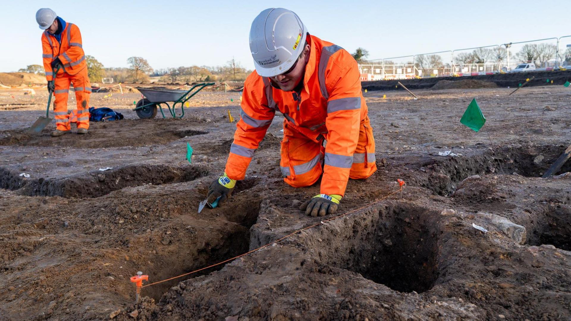 Two archaeologists wearing fluorescent orange jackets and trousers, one of them kneeling down, can be seen working on a site of bare earth with holes visible in the ground