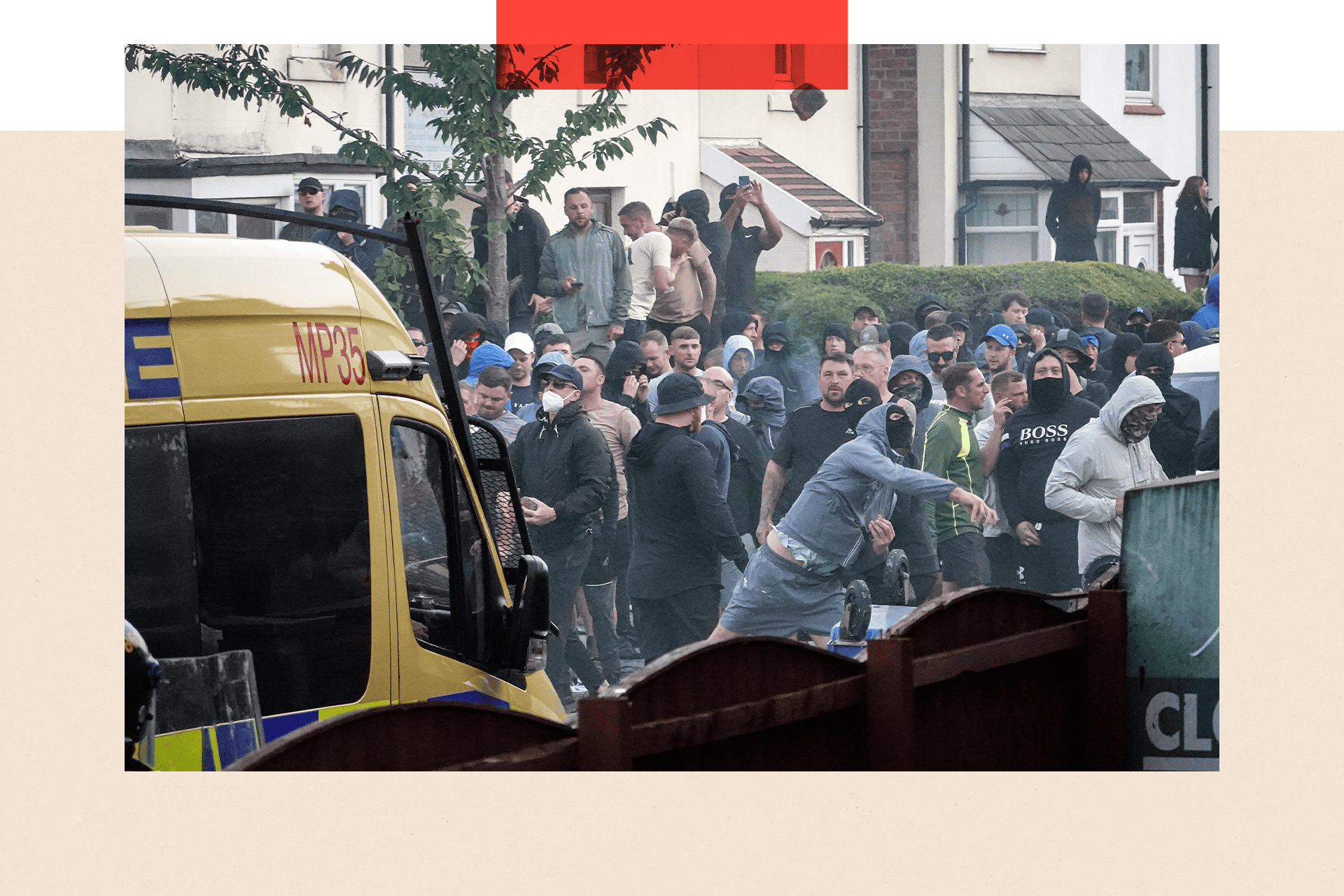 A crowd of male protesters behind a police van. One man in the foreground is wearing a black face covering and blue tracksuit top and shorts. He appears to be hurling something.