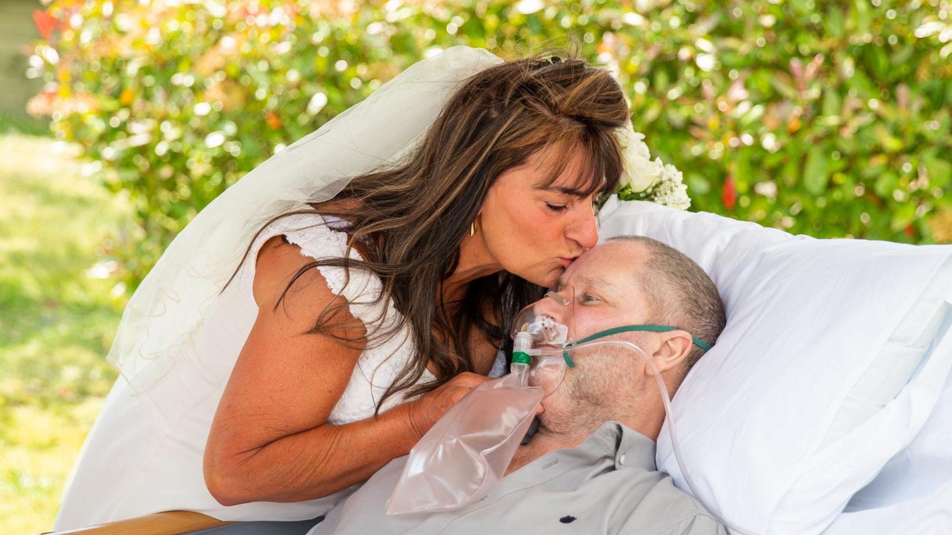 Annette Carter dressed in a wedding dress and veil. She is leaning down to kiss Sean Carter, who is wearing an oxygen mask, on the forehead