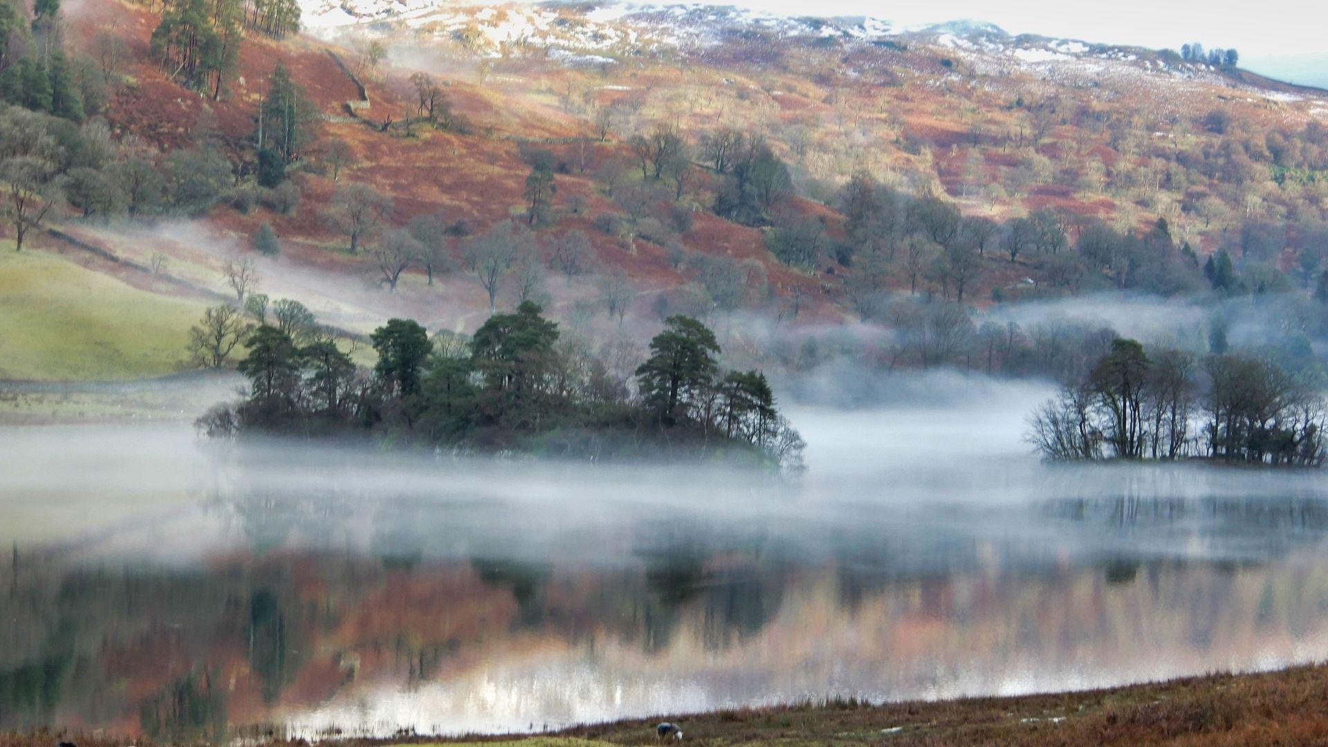 An island with a dozen or so green trees sits in the middle of a lake shrouded in a low thin mist, giving an ethereal quality. The hills beyond are brown and dark green with white patches of snow at the top. The island, hills and mist are all reflected in the serene and smooth lake surface.