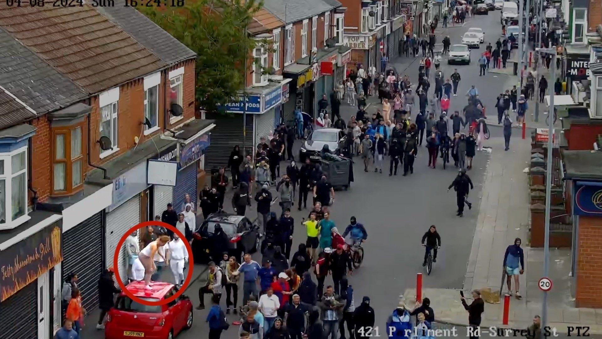 A mob of people standing in the road. Megan Davison can be seen circled in red standing on top of a red car in the bottom-left corner of the picture.