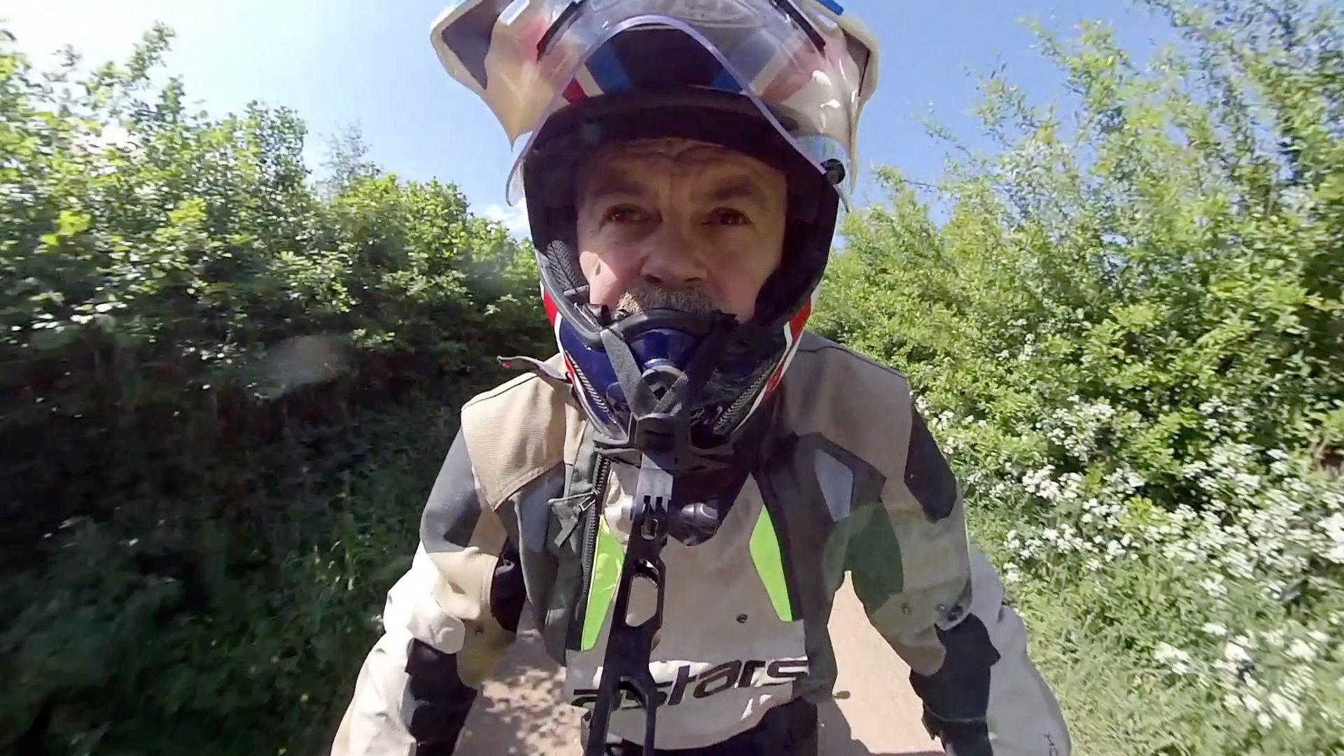 A close-up of a man on a motorcycle wearing a helmet, as well as a beige, grey and white motorcycle jacket. He is riding down a narrow path surrounded by bushes and white flowers.