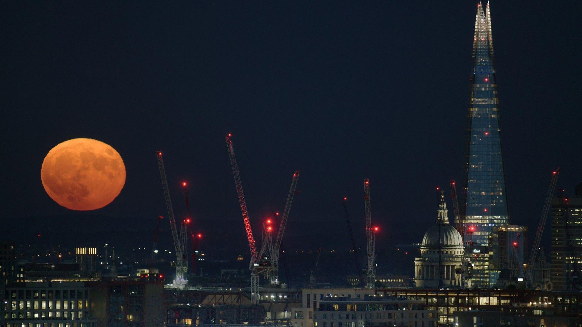 Full Strawberry Moon sitting low in the night sky behind London skyline including St Paul's Cathedral, the Shard skyscraper, building cranes and office buildings