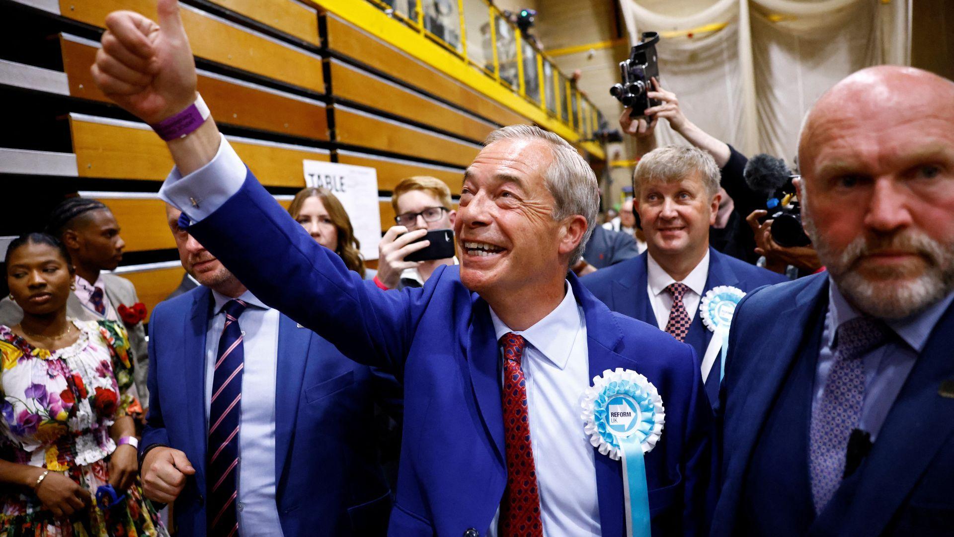 A group of people at an election count. In the middle is Nigel Farage with his thumb in the air.