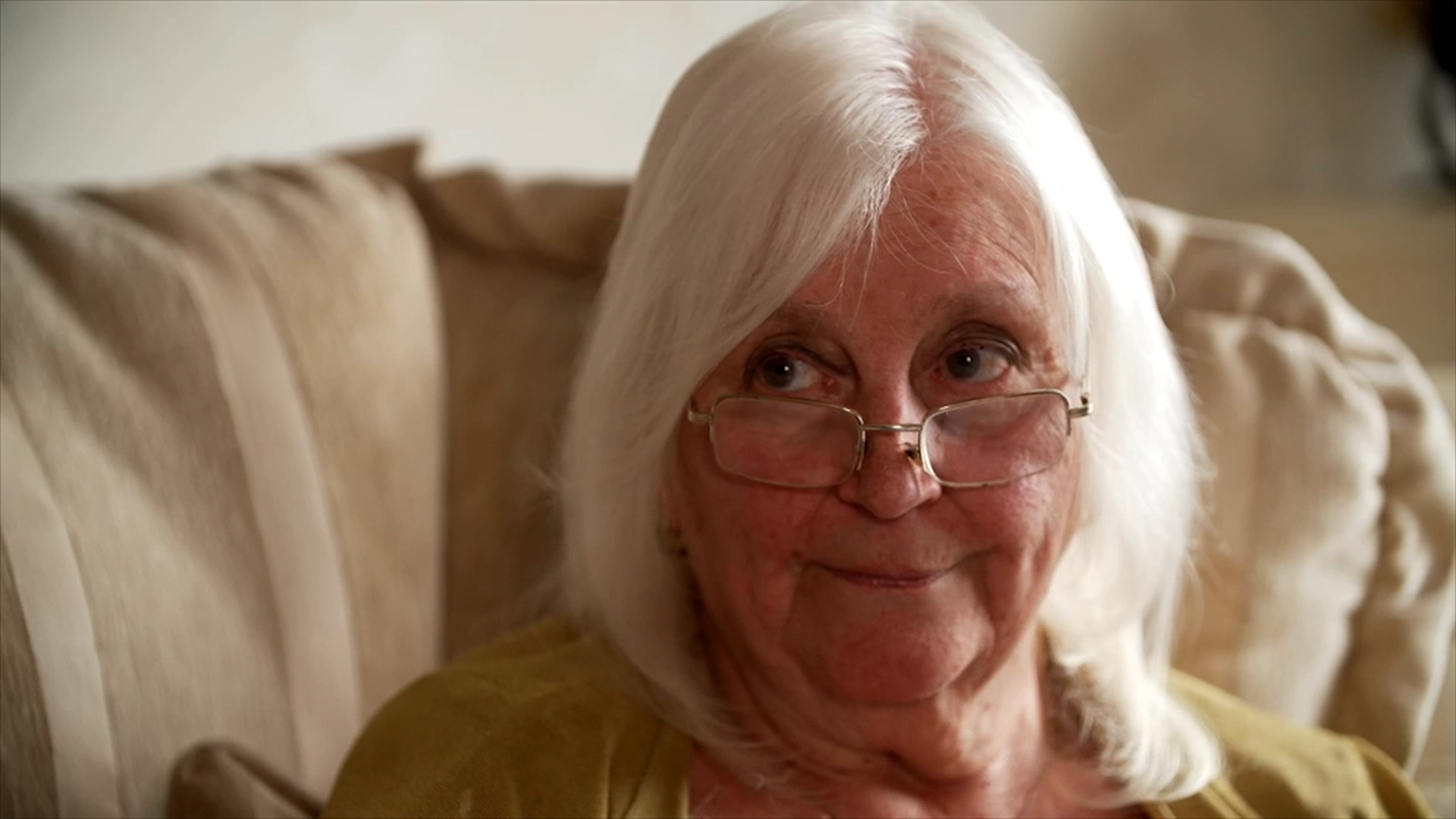 Sue is sitting on her cream-coloured sofa. She has white shoulder length hair and a pair of glasses perched on her nose. She is wearing a mustard top.