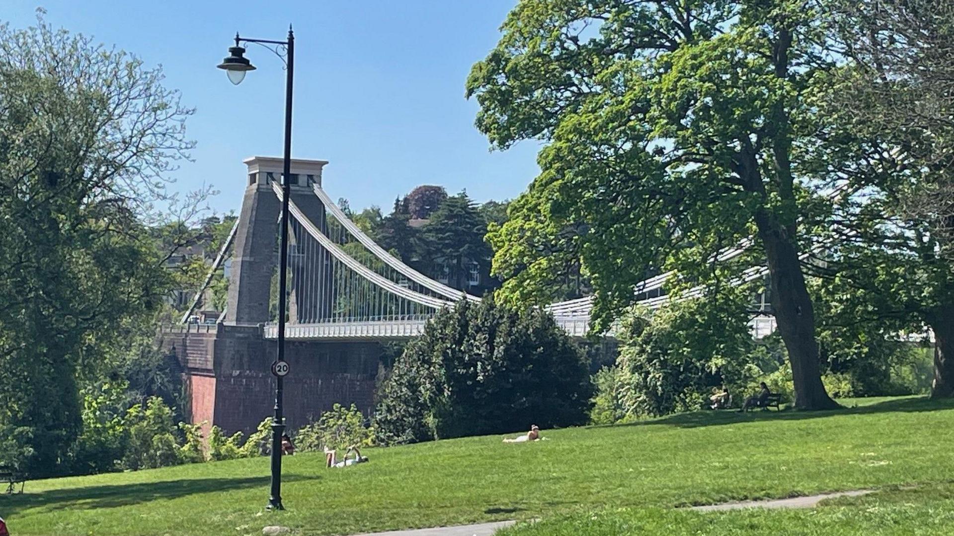 Two people are seen in the distance sunbathing by the Clifton Suspension Bridge on a sunny day