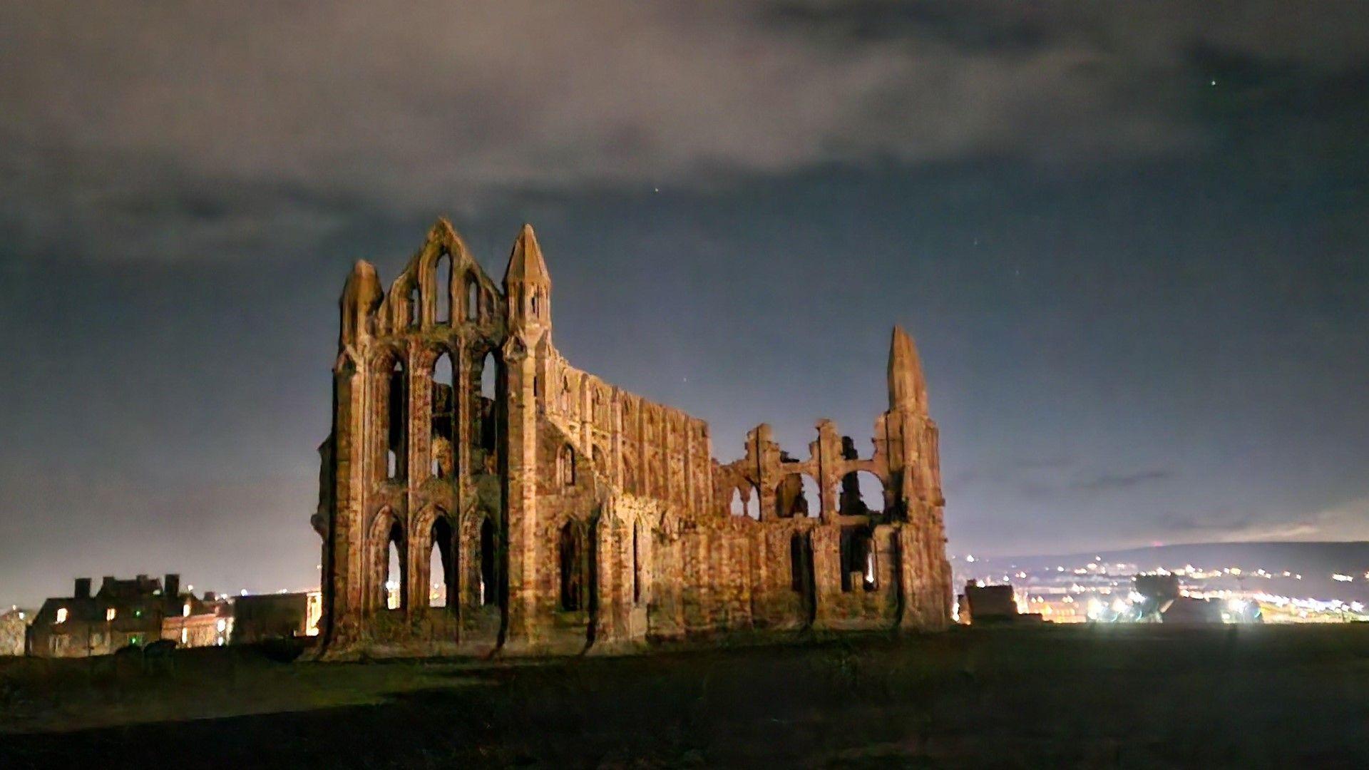 Whitby Abbey in foreground, illuminated. Clear spells in the night sky in the background