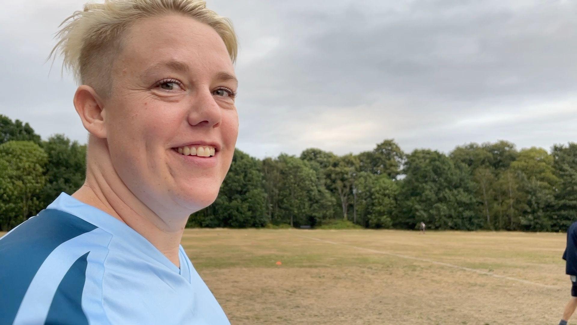Gelske Huyer has short blonde hair which is shaved at the sides. She's wearing a light-blue top and is standing on Wandsworth Common (a dry grassy field with trees in the background). She's smiling.