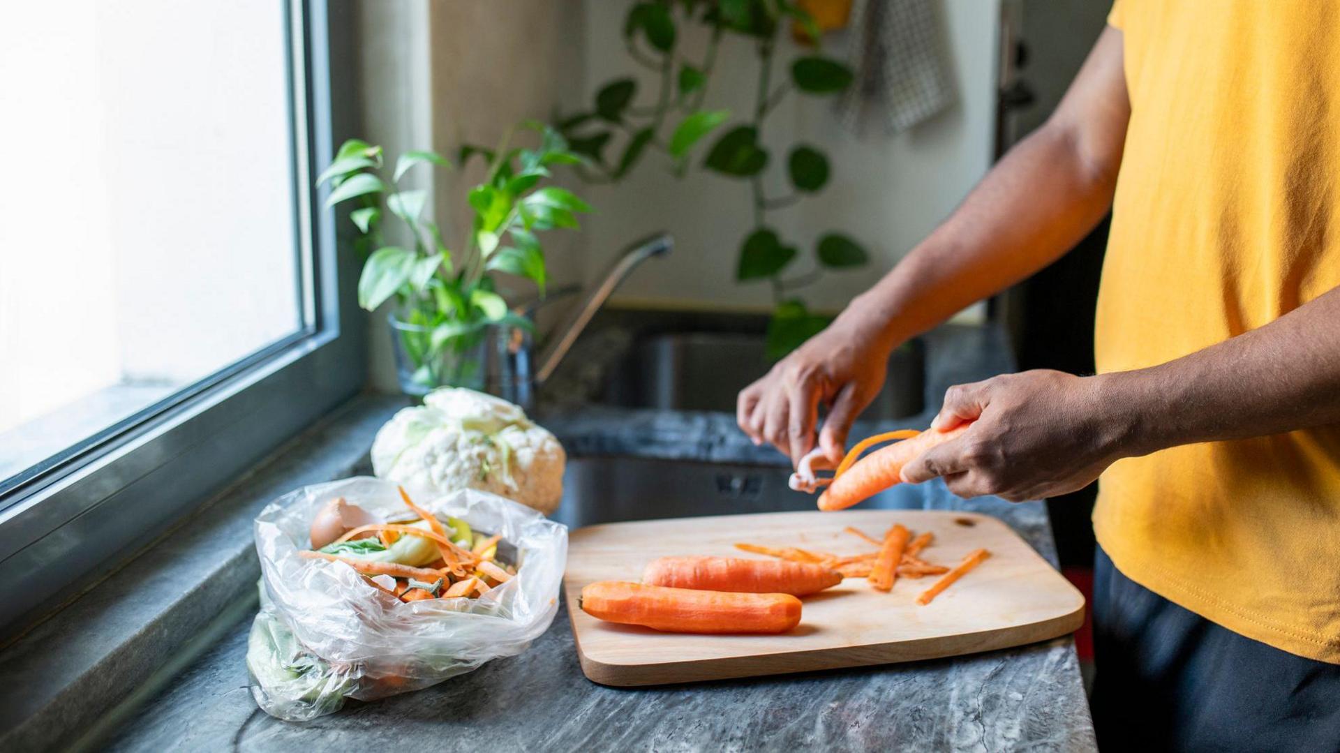 A person peels carrots over a wooden chopping board. They have a bag next to them with the peelings in and with other food waste. In the background there is a sink and plants.