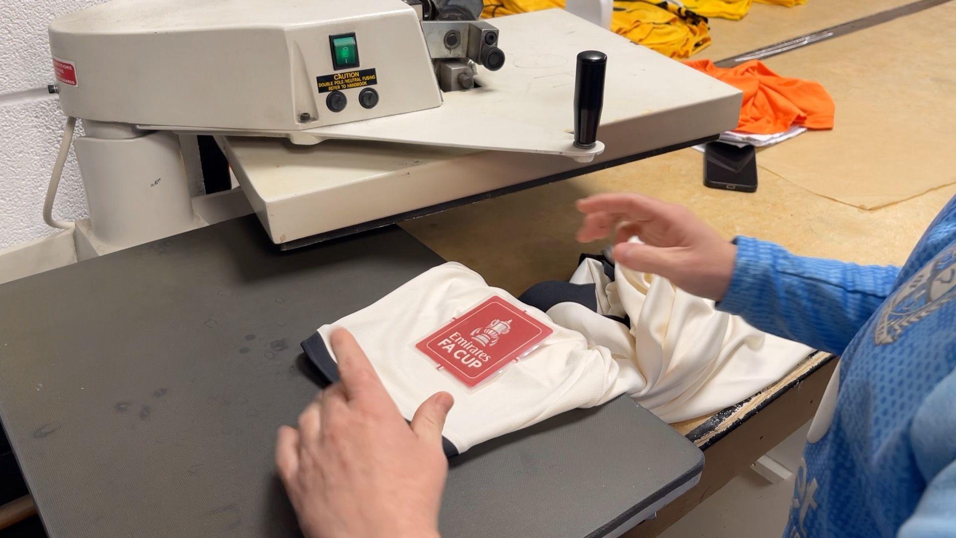 A red FA Cup patch being applied to a white Port Vale home shirt. 