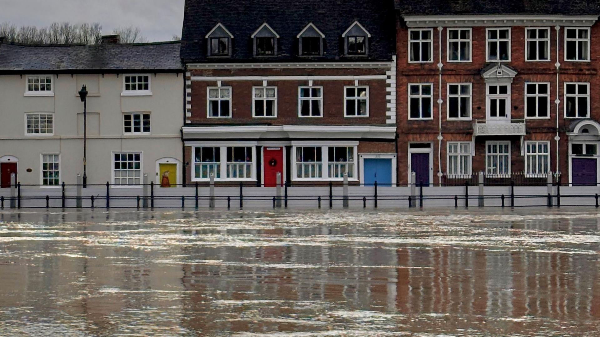 The River Severn, in flood, passes through Bewdley, with grey flood defences in operation and buildings behinds. Two are red-brick with white window frames, and the third is an olive colour with a yellow door