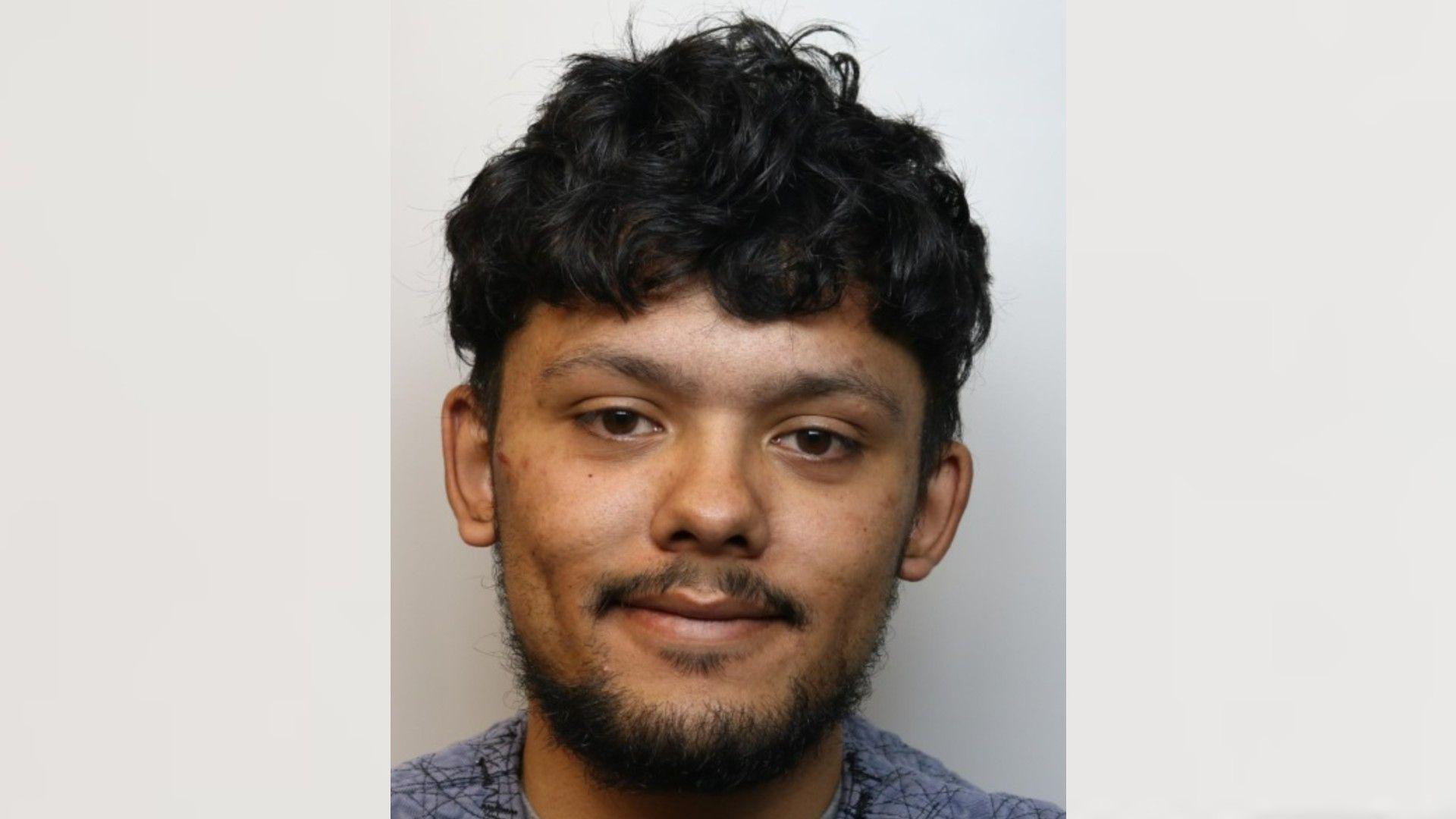 A custody mugshot of Claudio Stroud. He is wearing a grey jumper and is smiling at the camera. He has a dark beard and moustache and dark hair