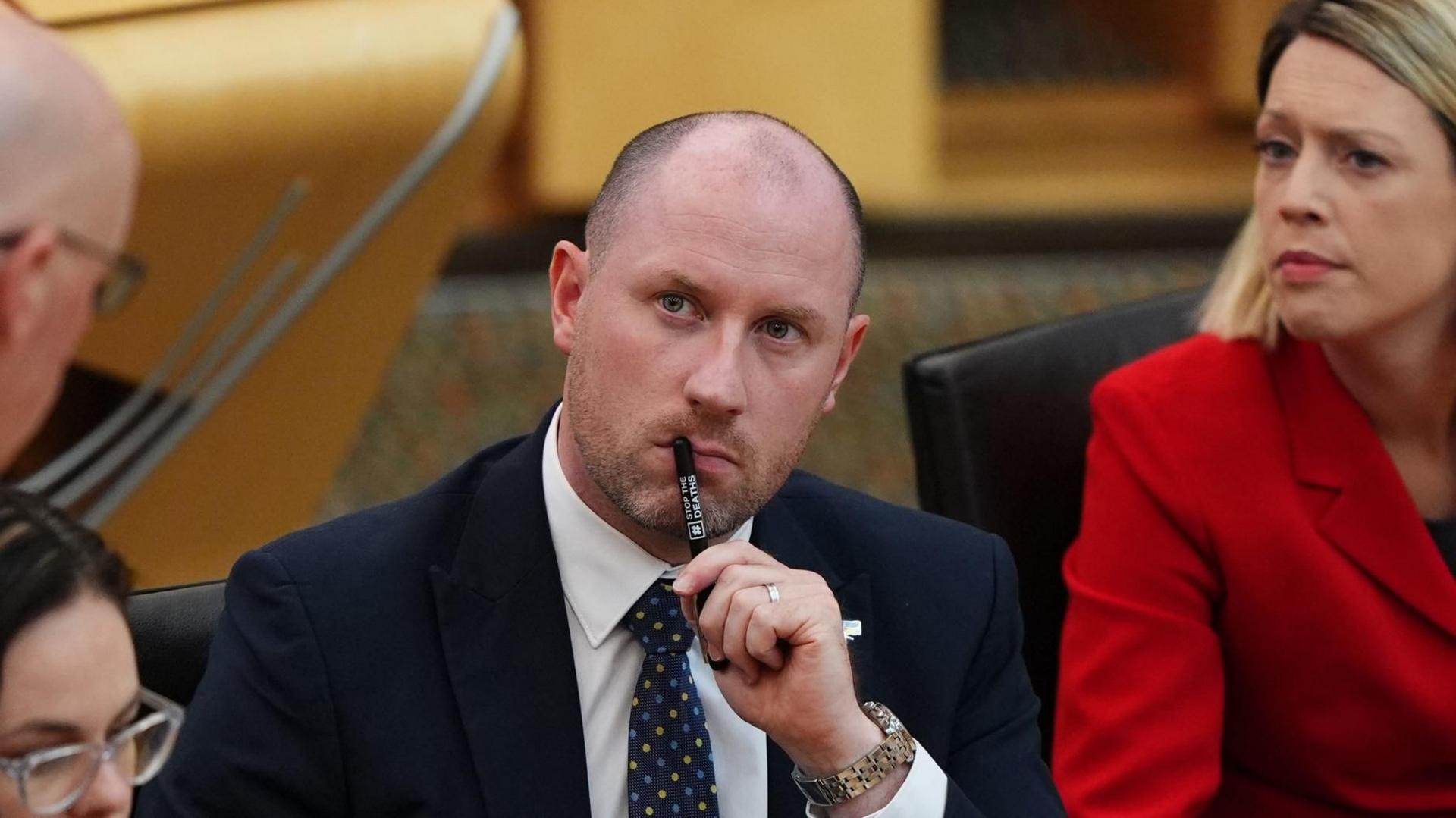 Neil Gray sitting listening in the Holyrood chamber. He is flanked by two female MSPs and is holding a pen to his lips while he listens. 