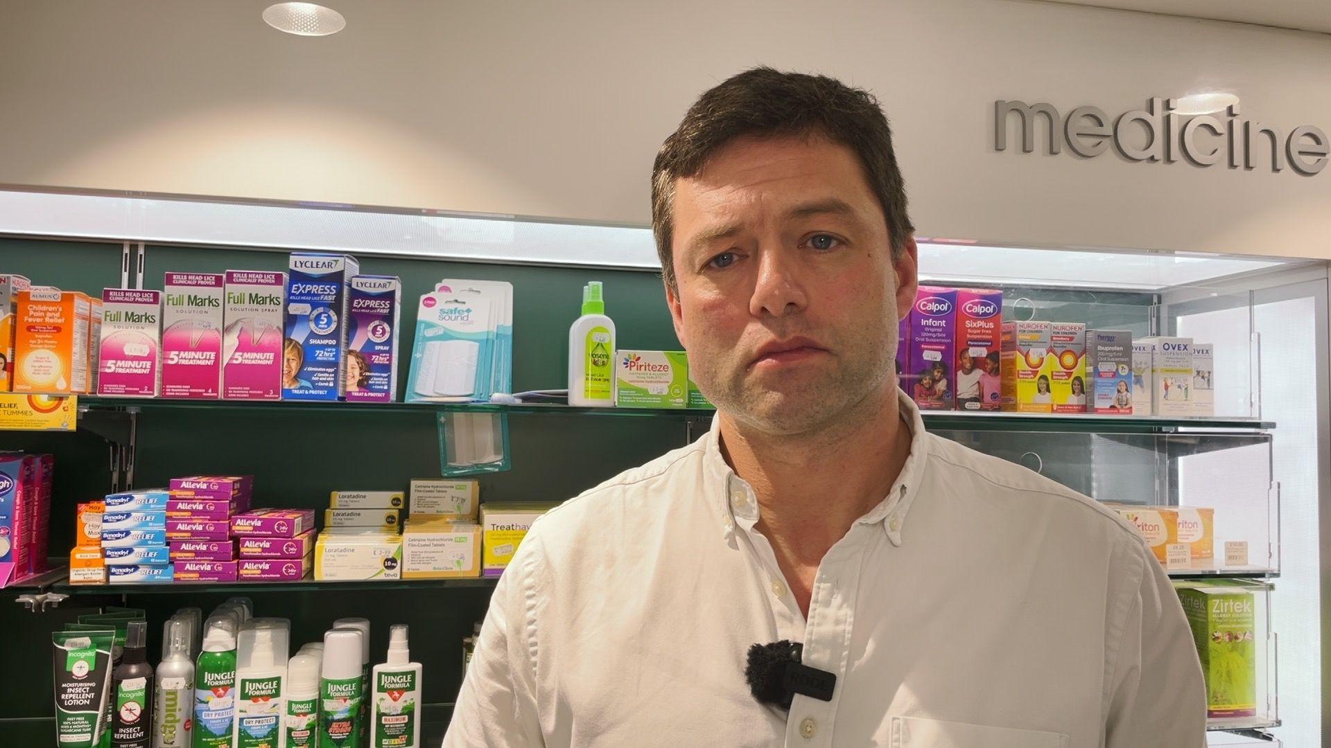 Chris Coghlan MP wearing a shite shirt and standing in a pharmacy with shelves of medicine behind him.