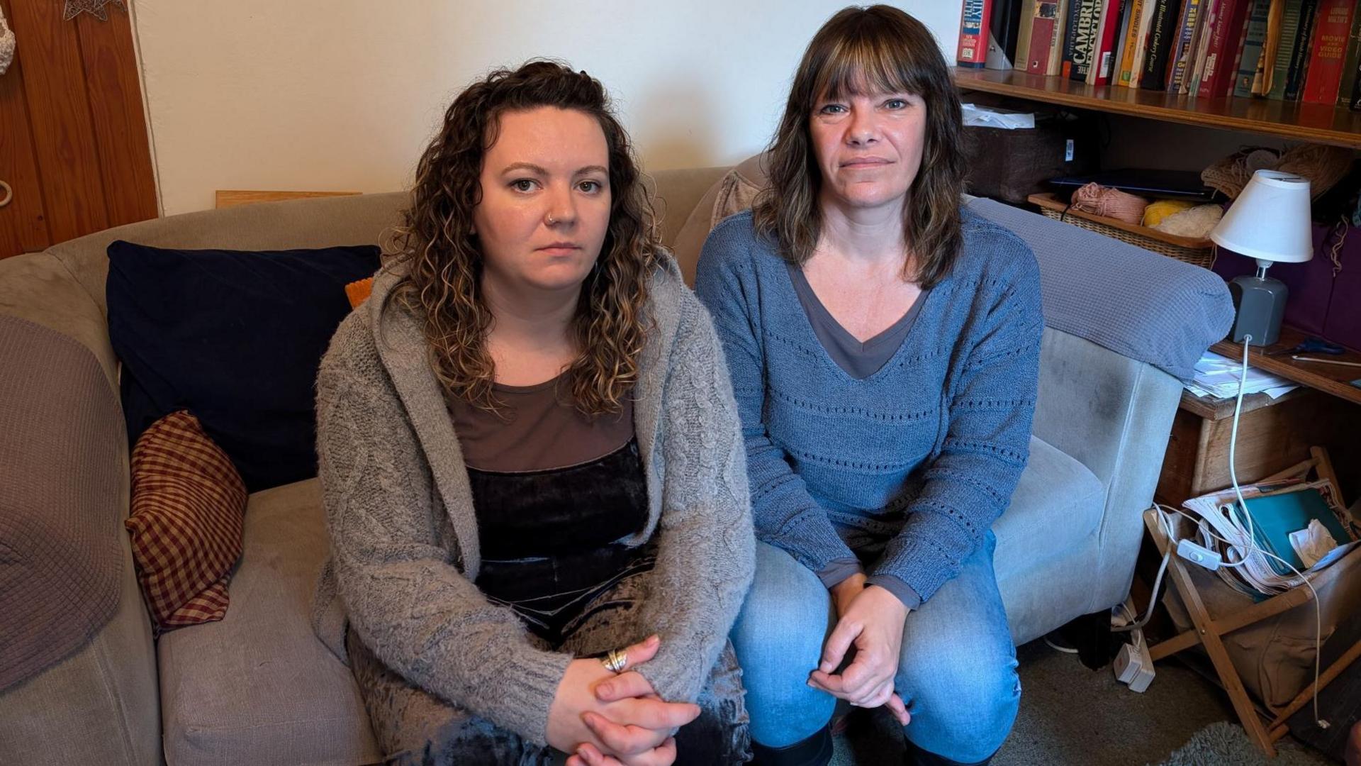 A picture of Juliet Rimer with long, curly, brown hair and wearing a black top and grey cardigan. She is sat on a sofa next to a bookshelf alongside Kate Rimer, who has long, brown hair and is wearing jeans and a blue jumper. 