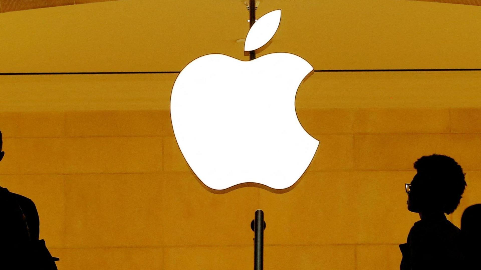 Apple logo glows in white on yellow background with shadows of people walking past