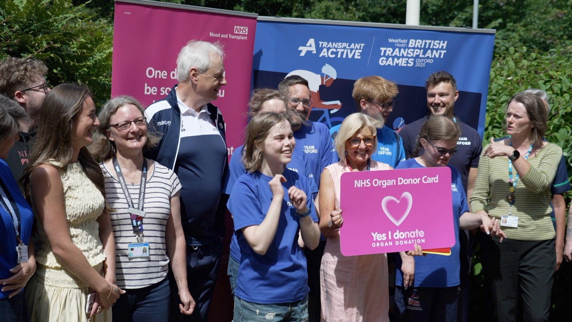 A group of people standing together, whilst one in the middle holds a large pink organ donation card.