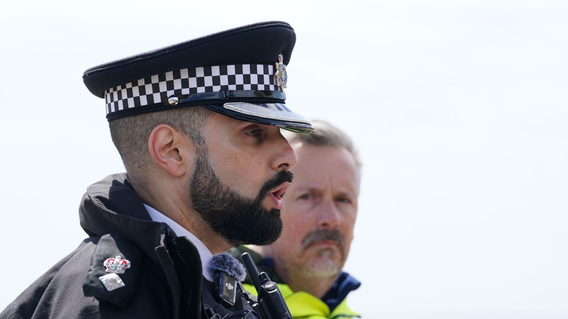 Profil anësor i një oficeri policie mashkull me mjekër të zezë. Në të majtë të tij është një burrë me një xhaketë me dukshmëri të lartë.
