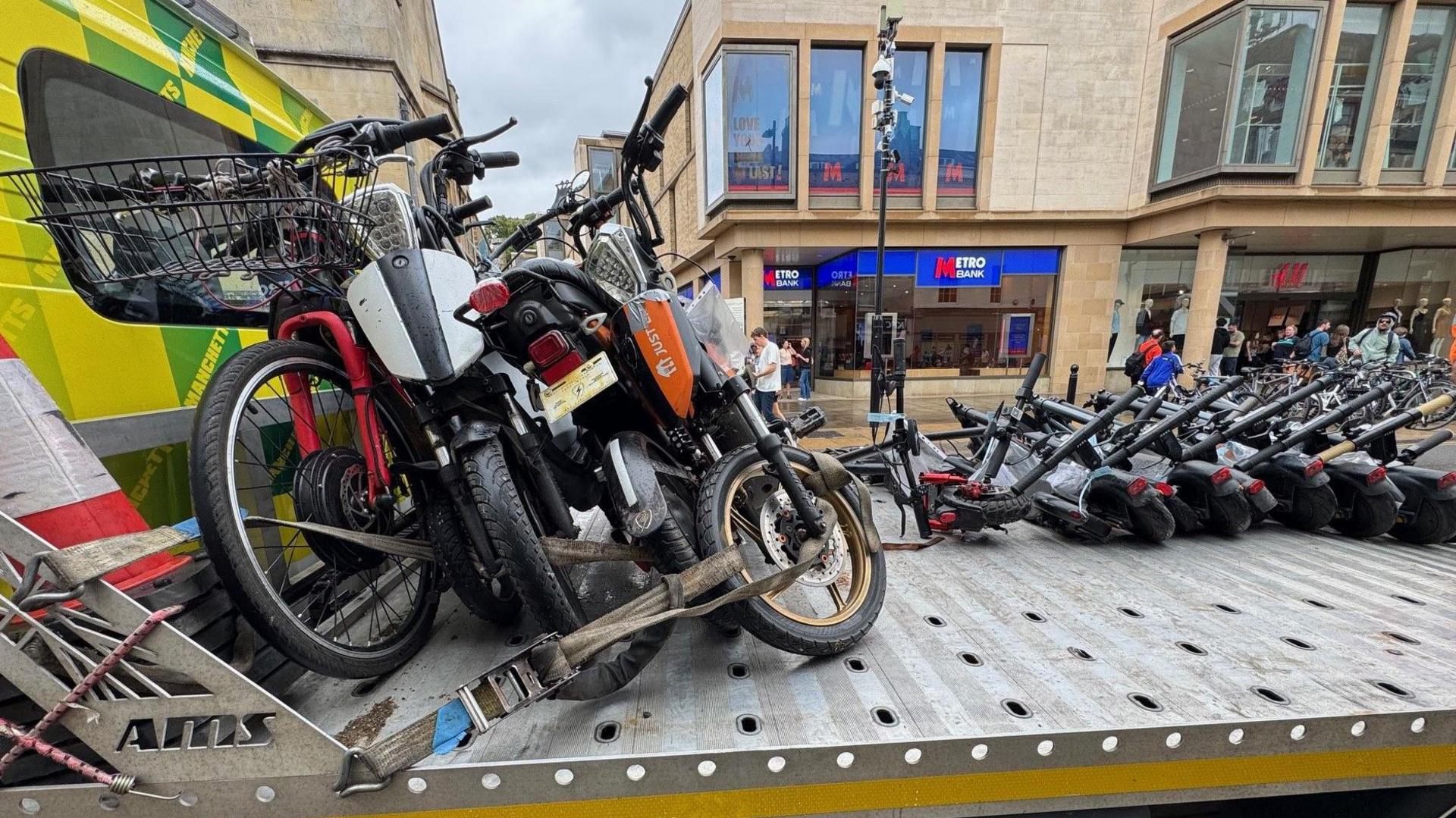 A flatbed truck with no edges, loaded with confiscated vehicles. There are about four e-bikes on the left, strapped on to the truck, and about six black e-scooters on the right. Behind the truck are Metro Bank and H&M.