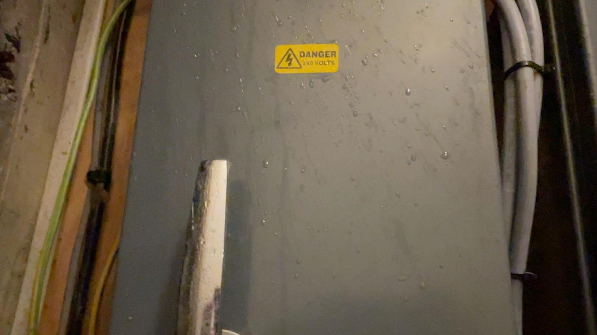 A grey electricals box is in the middle of the image. A yellow sticker is in the middle which says DANGER on it and has a sign. There are wires either side of the box.