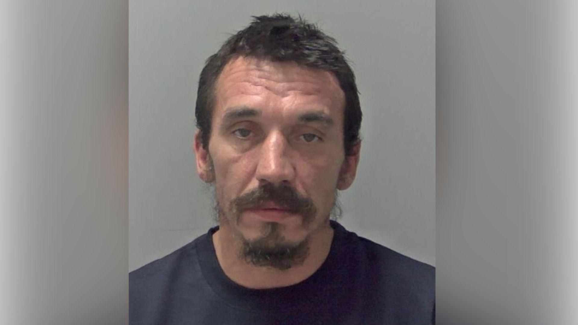 Fred Lamb with short dark hair and beard wearing a dark top in a police custody photo.