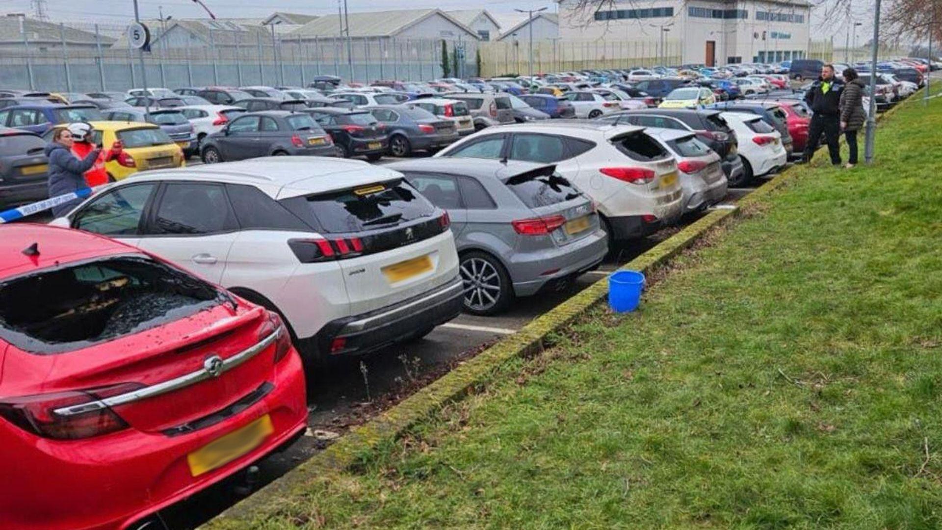 A car park with dozens of cars, several of which have smashed windows. Police tape is visible in front of some of them, and the cars back on to a grass verge. 