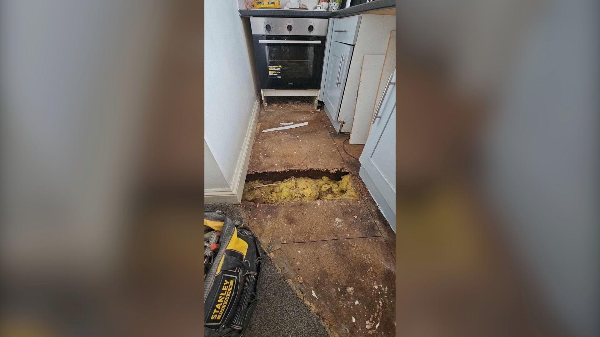 A narrow galley kitchen with an oven and white kitchen cupboards. The floor is bare boards and there is a large hole in the middle where you can see insulation. There is a black tool box in the foreground.