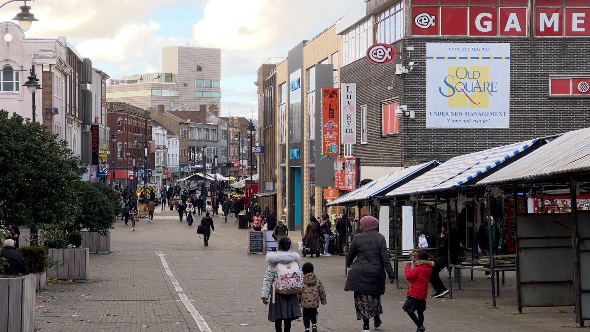 A town centre with market stalls, shops and people walking down the street. The outdoor market stalls are empty. There is a woman and three children at the front of the image, all walking down the street.