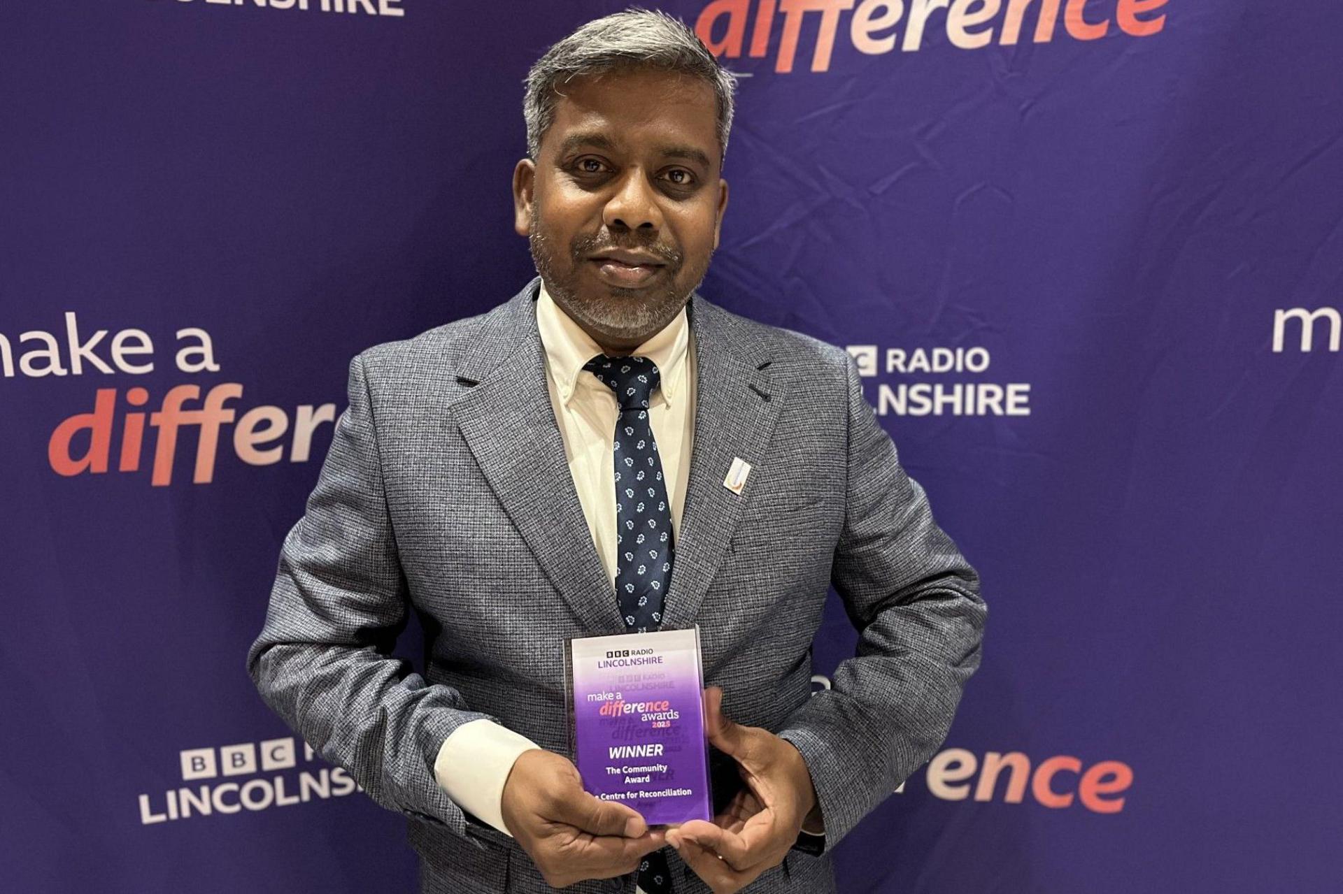 A man wearing a grey suit with a white shirt and a blue patterned tie holds a rectangular-shaped award, made of glass coloured purple and white, with writing that reads: "BBC Radio Lincolnshire Make a Difference Awards, Winner".