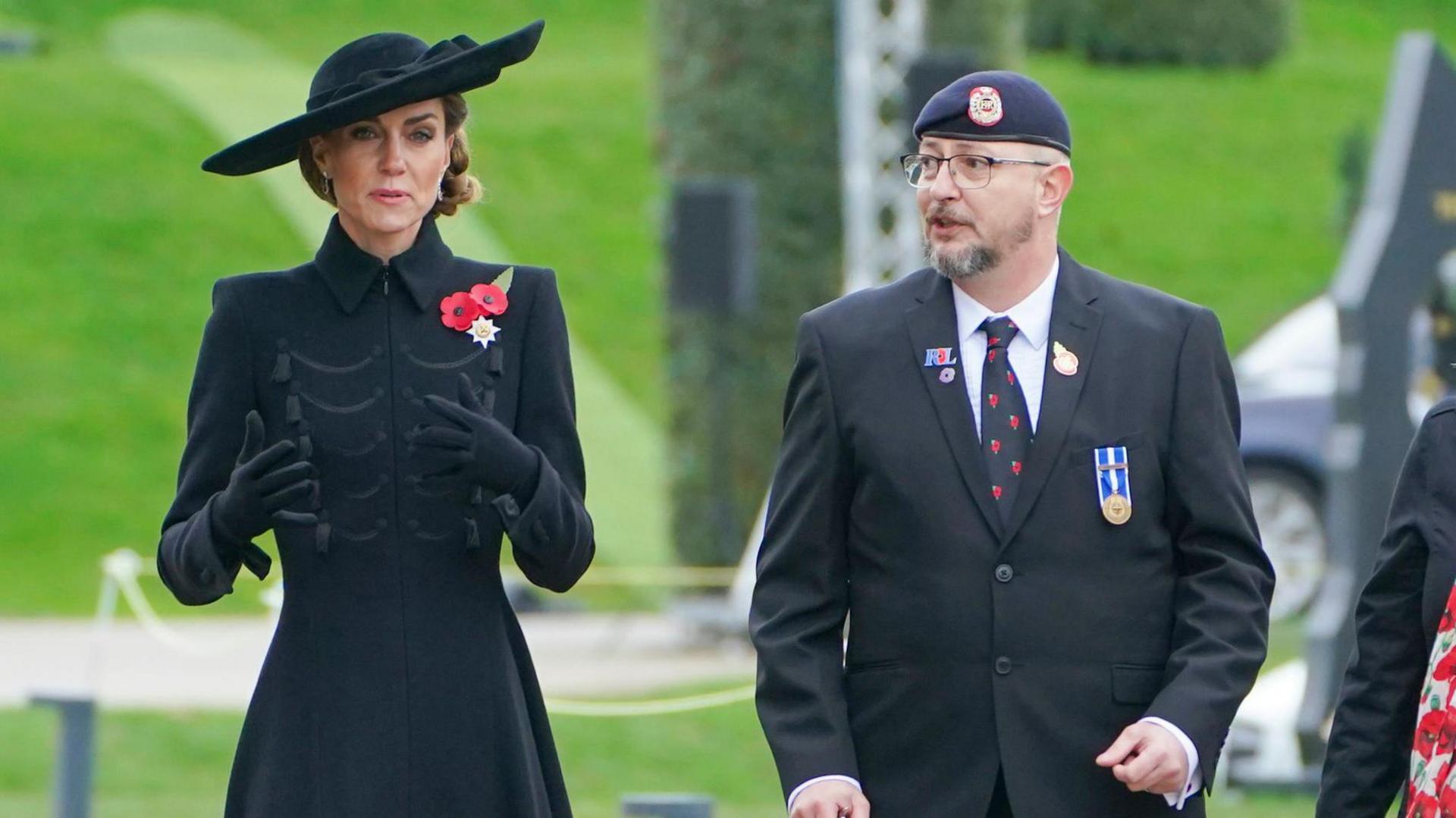The Princess of Wales is walking beside a man in a black suit and navy beret. She is dressed in a large black hat, black gloves, a long black coat with two red poppies on her left chest and her brown hair is tied up. The man is wearing a white shirt, black tie decorated with poppies, a medal and three pin badges, one of which is shaped like a poppy. Grass, a headge and a guidance rope can be seen blurred out behind them.
