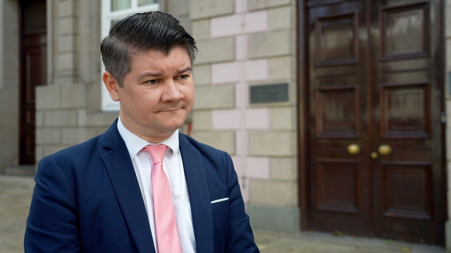 Deputy Sam Mézec stood outside the States Assembly building. He has short dark brown hair and is wearing a white shirt with a blue jacket and pink tie.